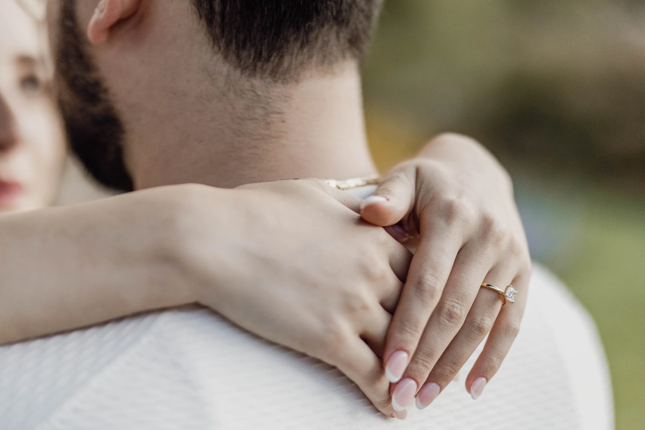 A woman with long nails and a ring hugs a man with a beard, with focus on their embrace and the man's neck.
