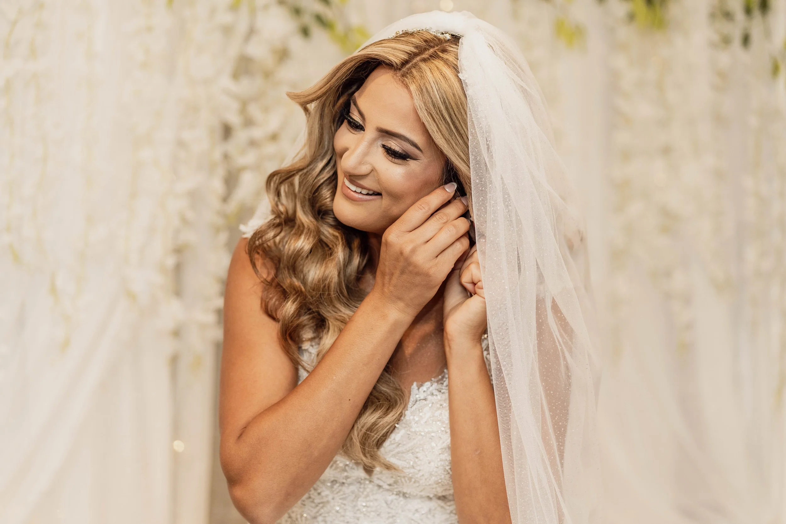 A smiling bride with long wavy hair, wearing a lace wedding dress and a veil, is adjusting her earring in front of a floral backdrop.