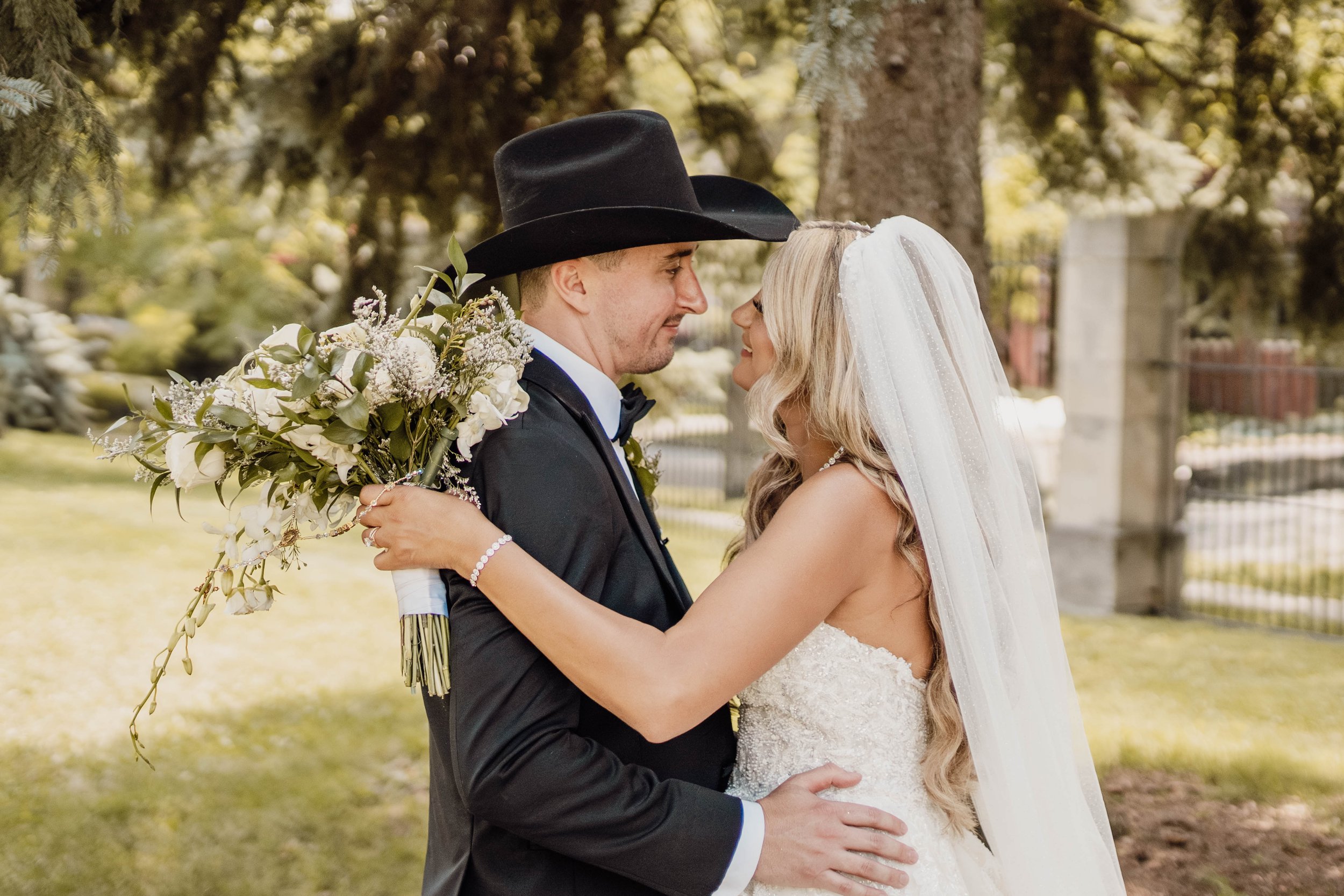 Bride and groom in wedding attire embracing outdoors, with the groom wearing a black cowboy hat and holding a bouquet of white and green flowers.