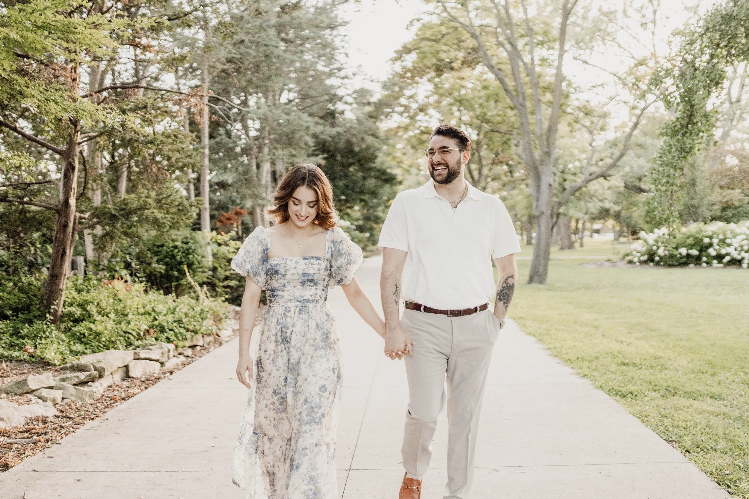 A couple holding hands and walking along a sidewalk in a park during daytime, smiling and enjoying each other's company, surrounded by trees and greenery.