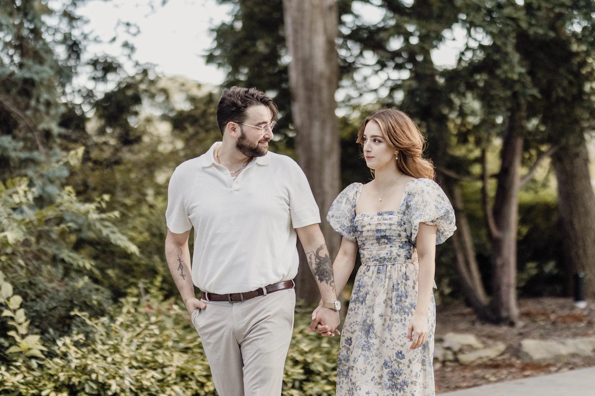 A couple holding hands and walking outdoors in a park surrounded by trees and foliage.