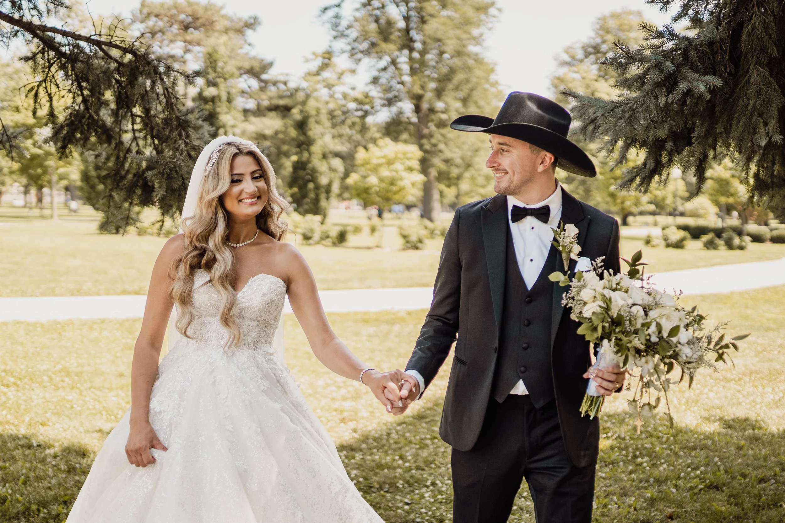 A bride and groom holding hands outdoors on a sunny day, with trees in the background. The bride is wearing a strapless white wedding gown and veil, and the groom is dressed in a black tuxedo with a cowboy hat, holding a bouquet of flowers.