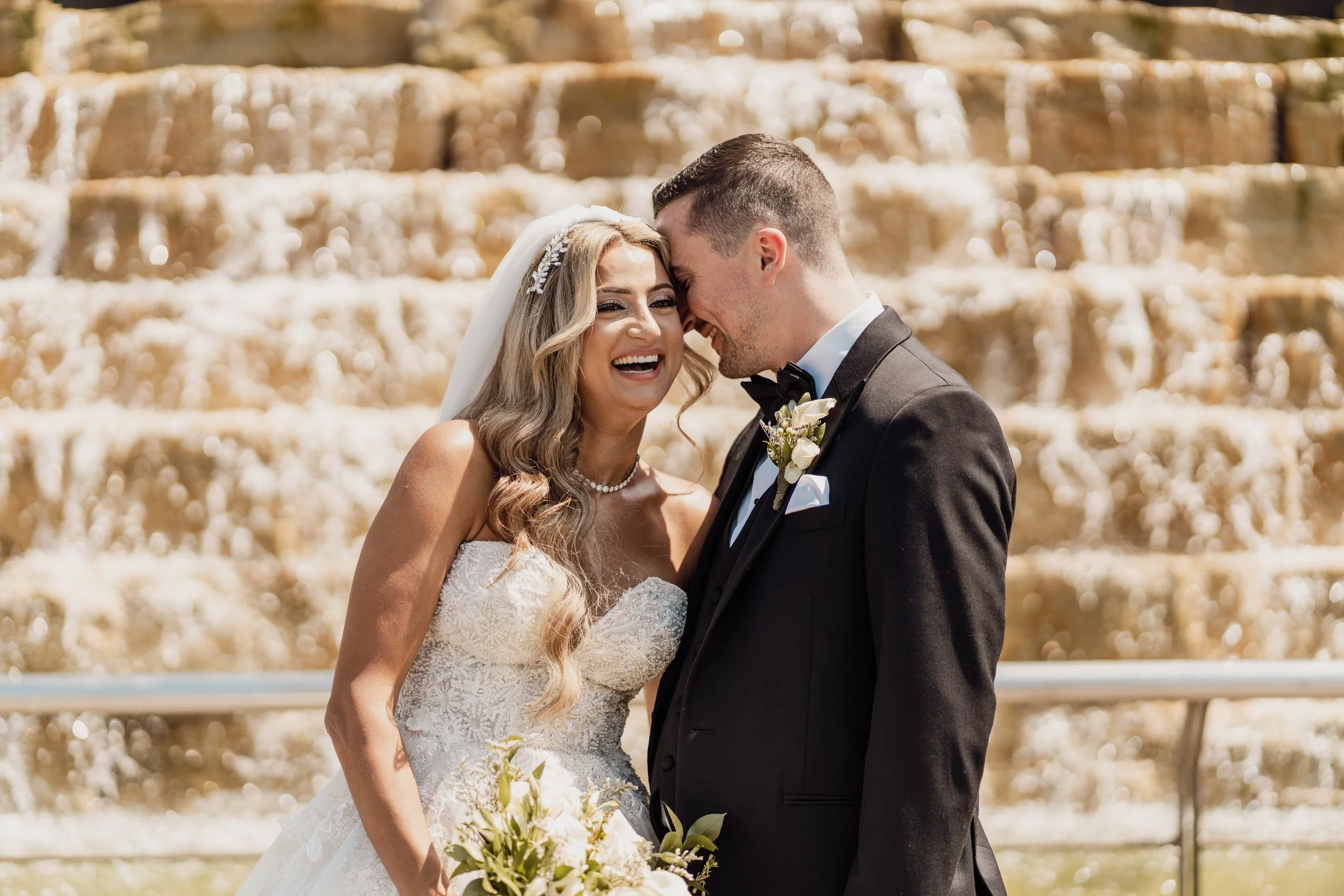 A bride and groom sharing a joyful moment during their wedding ceremony outdoors, with a cascading fountain in the background.