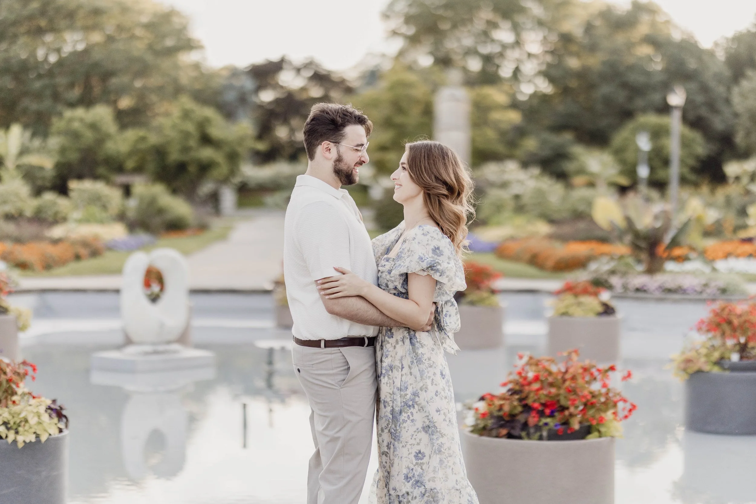 A couple stands close together, looking into each other's eyes and smiling, in a park with colorful flowers and greenery in the background.
