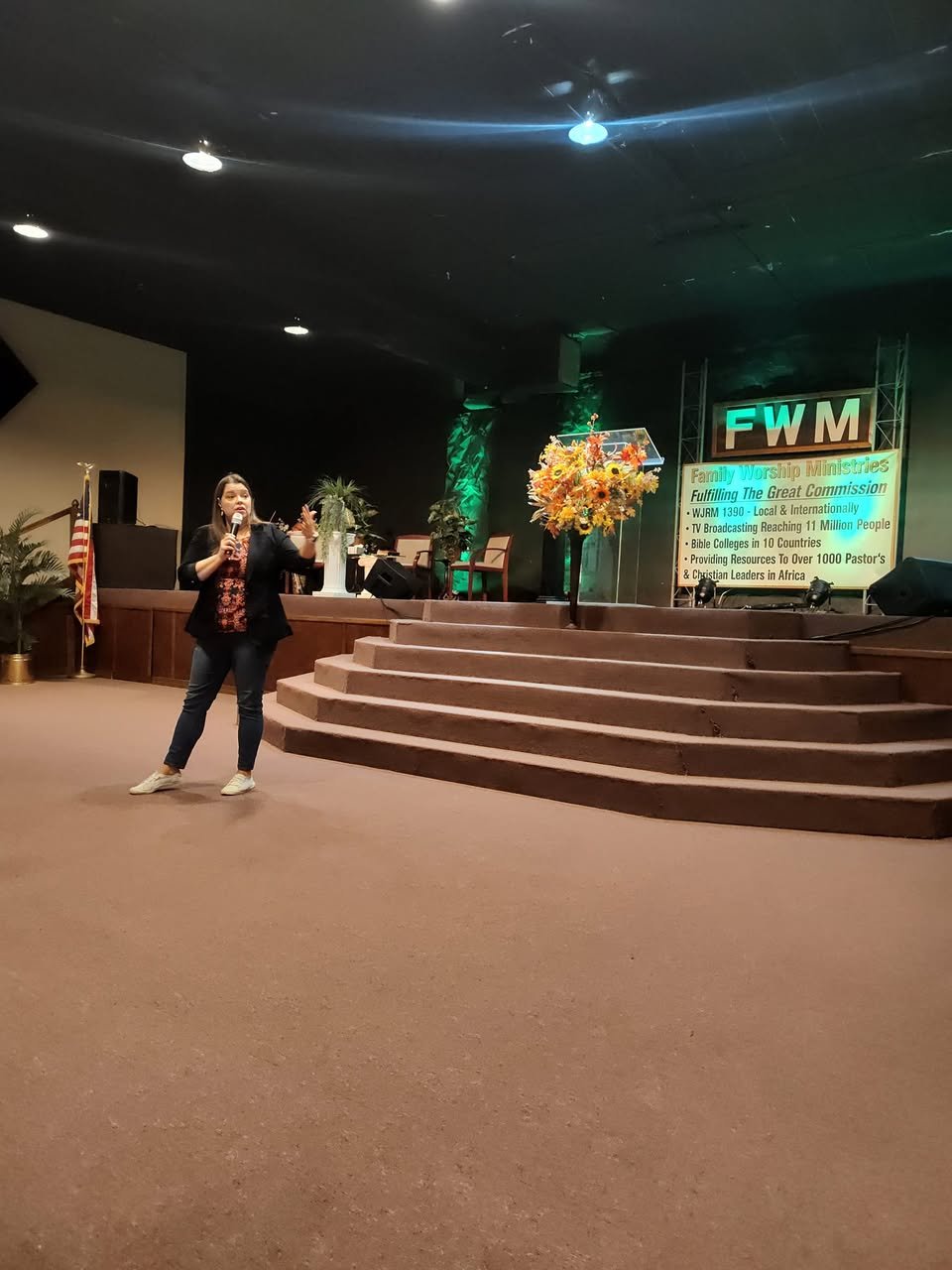 Danita Cummins speaking into a microphone on a church stage with a large flower arrangement and a digital sign in the background.