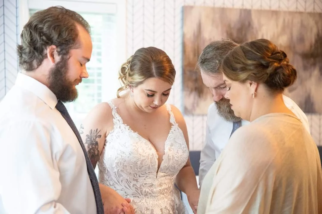 Danita Cummins praying with heads bowed, including a bride in a white lace wedding dress, during a wedding ceremony indoors.