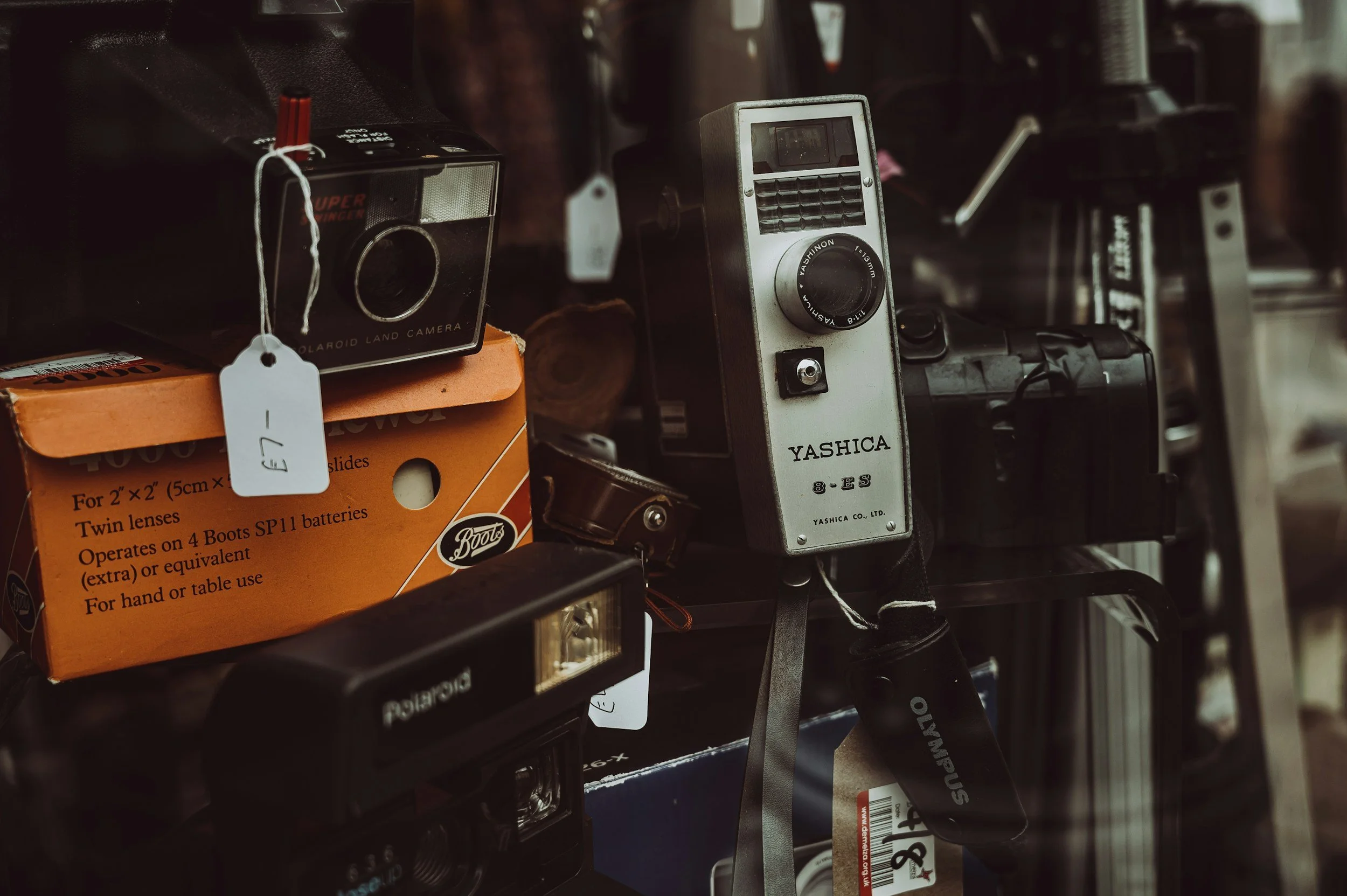 Collection of vintage cameras, including a Polaroid, Yashica, and Kodak, displayed on a store shelf with tags and packaging.