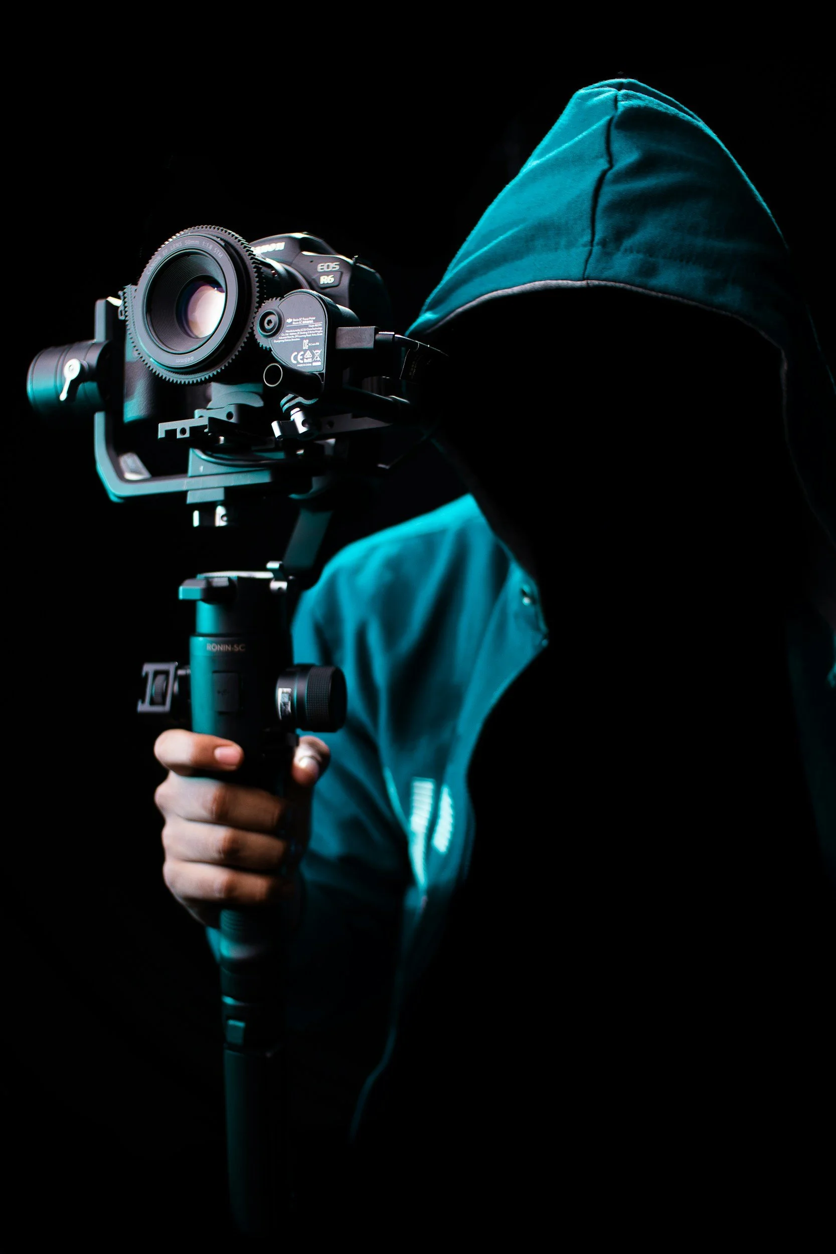 A person wearing a teal hoodie holding a professional camera on a stabilizer against a black background.