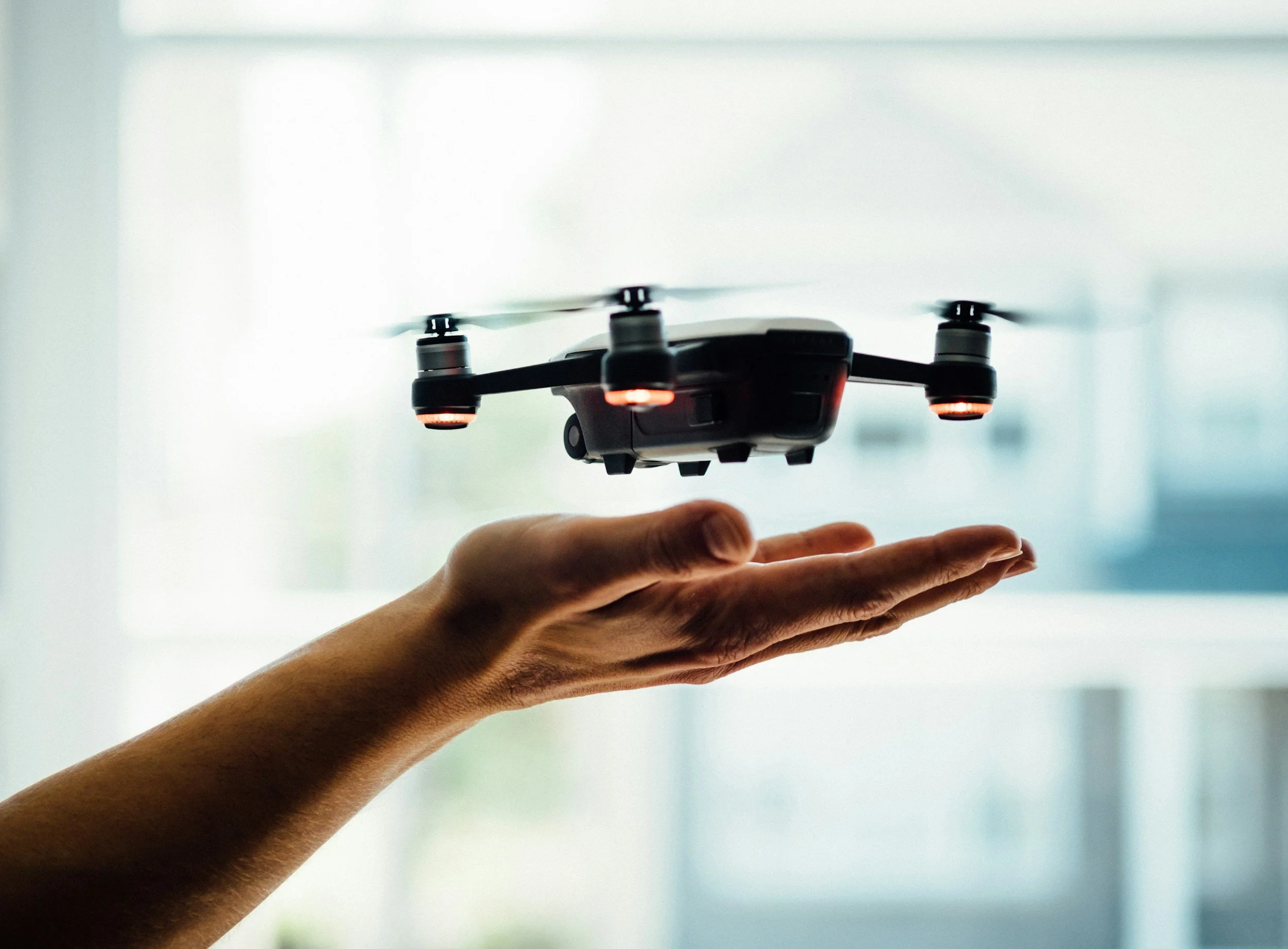 A person's hand is underneath a flying drone with red lights, set against a blurred background of windows and natural light.