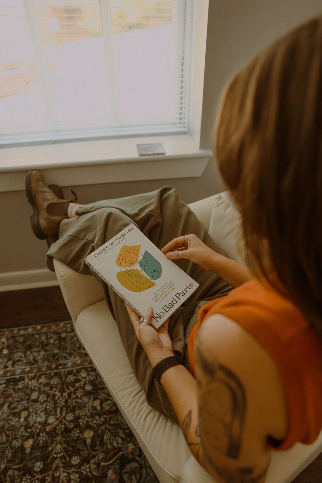 A woman with tattoos on her arm is sitting on a beige couch, reading a book titled 'No Bad Parts' by Richard C. Schwartz, PhD. She is wearing an orange sleeveless top and beige pants, with brown boots. She appears to be in a relaxed setting near a window with blinds, and a decorative rug is visible on the floor.