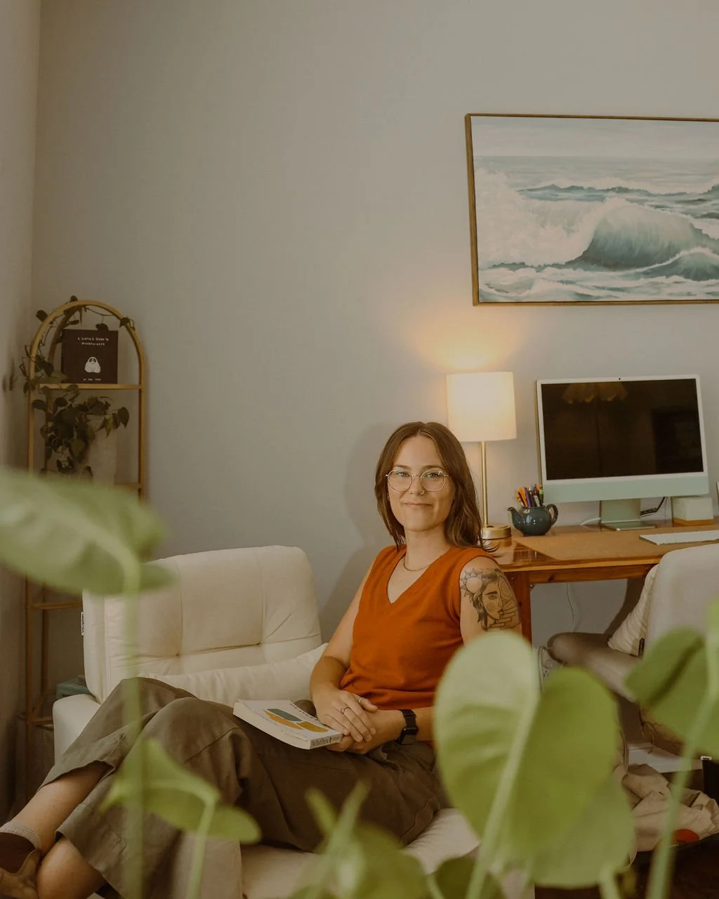 A woman with glasses and short brown hair, wearing an orange sleeveless top and brown pants, sitting on a white sofa in a cozy home office. She's holding a book and looking at the camera with a slight smile. In the background, there's a wooden desk with a computer, a lamp, and office supplies, along with a wall painting of ocean waves.