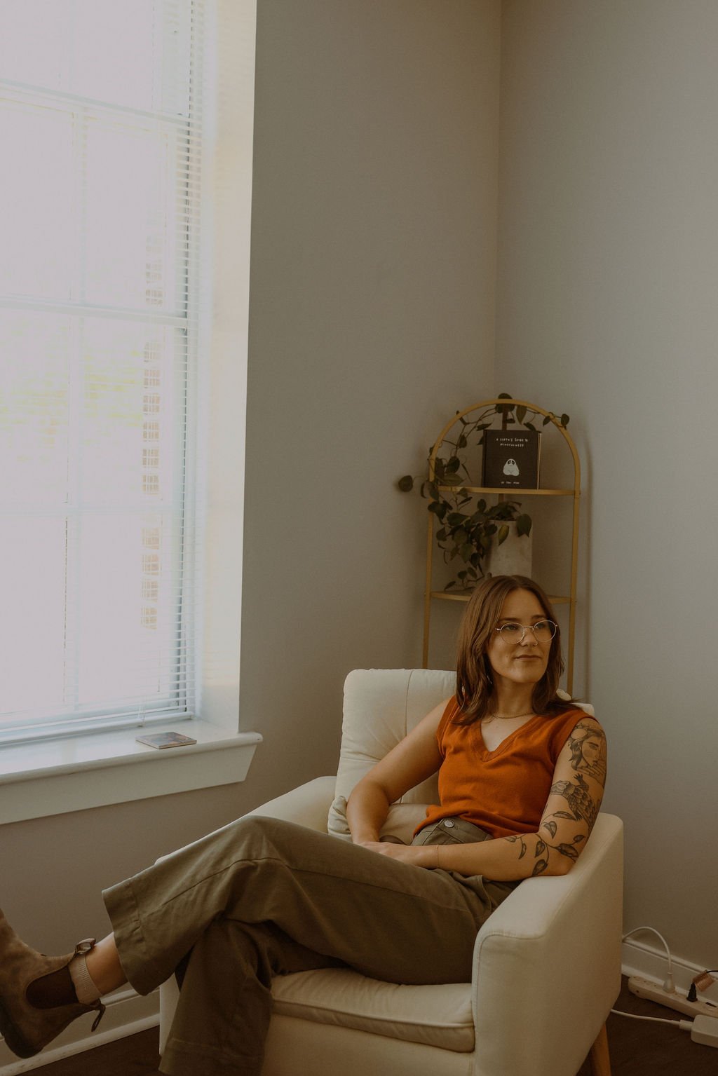 Woman sitting on a white armchair near a window with blinds, wearing glasses, an orange sleeveless top, and brown pants, with tattoos on her left arm, in a room with a white wall and a small gold shelving unit with plants and a book.