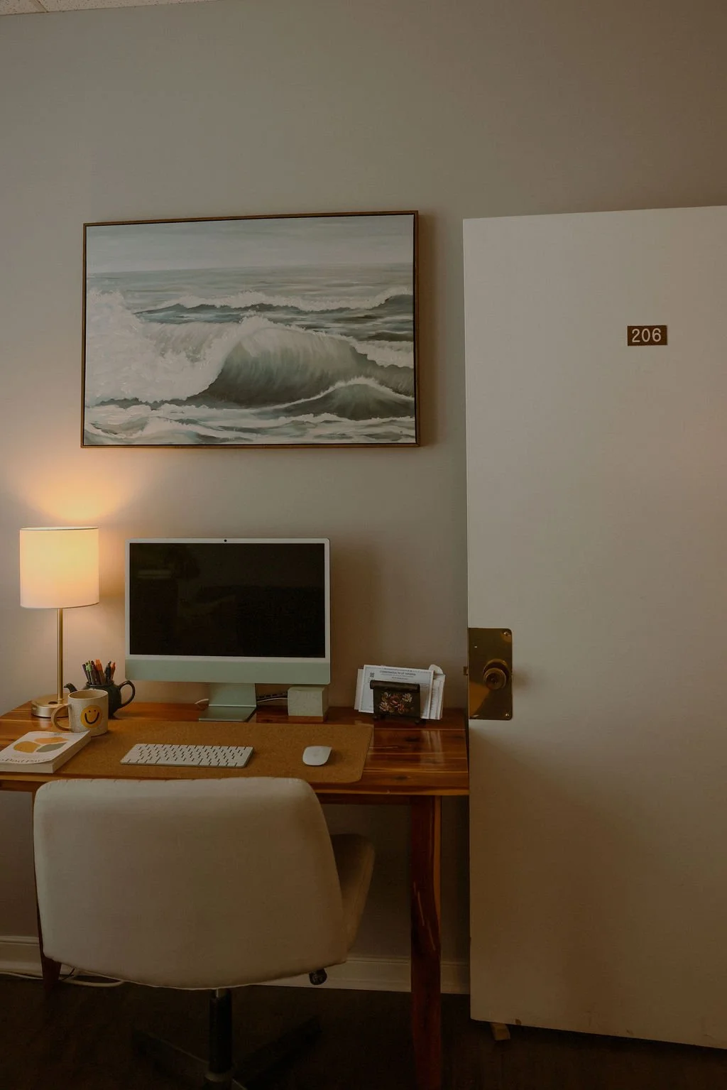 A home office with a wooden desk, a computer, a desk lamp, a coffee mug, and a painting of ocean waves on the wall.