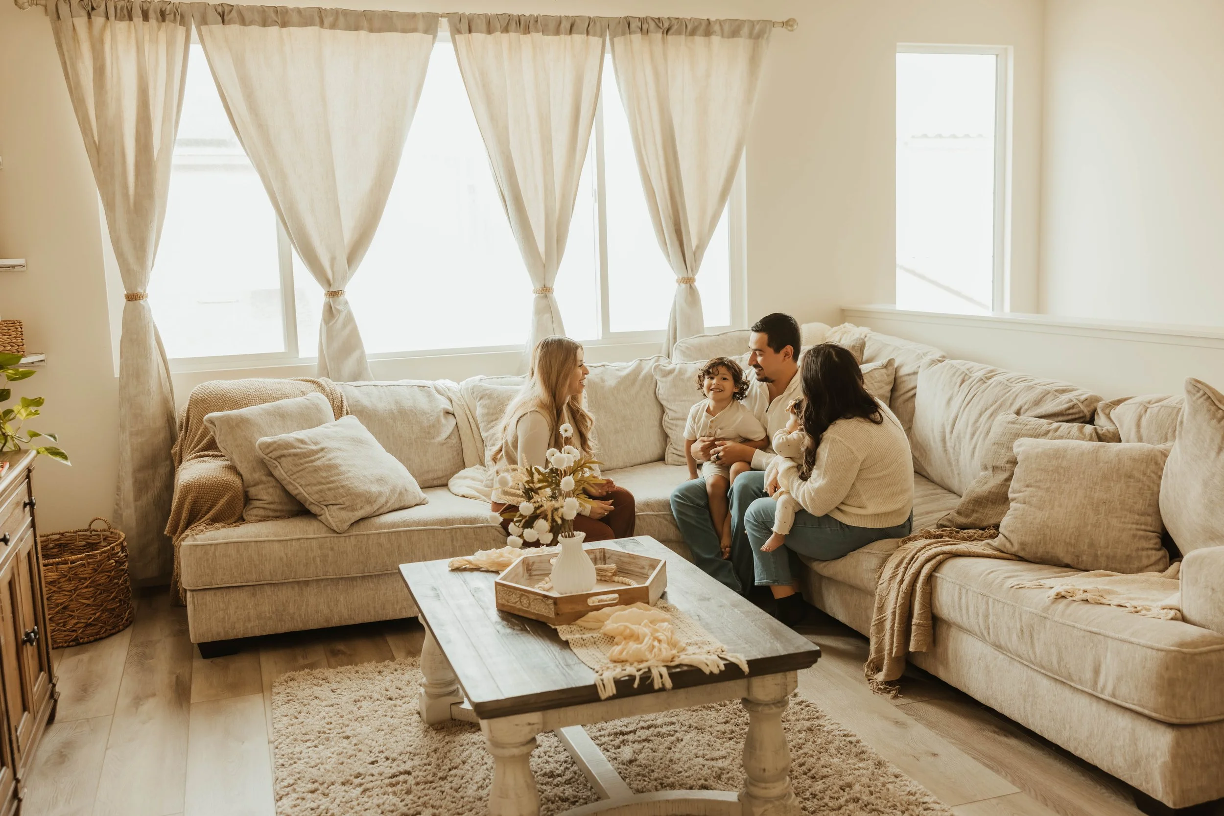 A family of four sitting on a beige sectional sofa in a bright living room, smiling and interacting near large windows with beige curtains. A wooden coffee table with a tray, a vase, and a blanket is in the foreground. The room has light-colored walls, wooden flooring, and cozy decor.