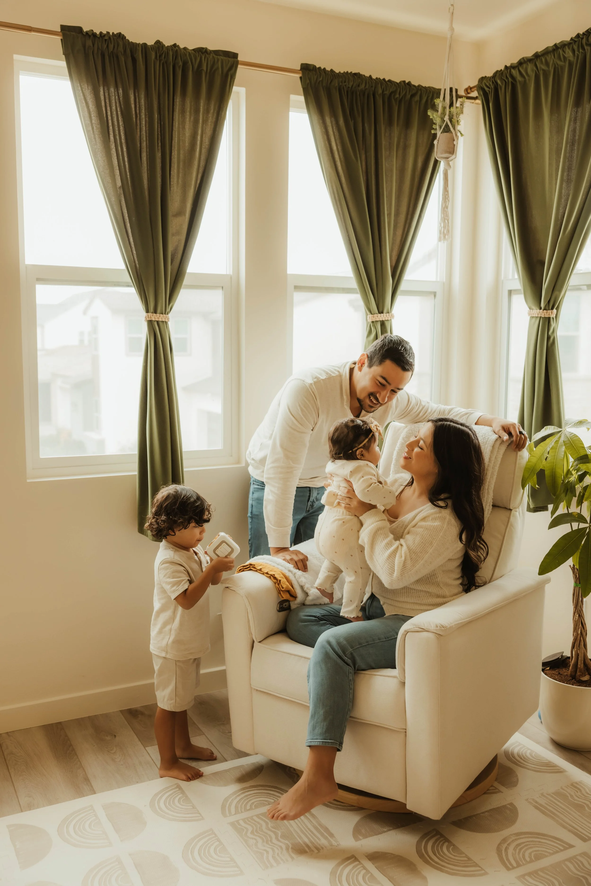 A family of five enjoying time together in a bright living room with large windows and green curtains. The parents are sitting on a white armchair, with the mother holding a toddler, and the father smiling beside her. Two young children are nearby, one holding a toy and the other standing, looking at the family.