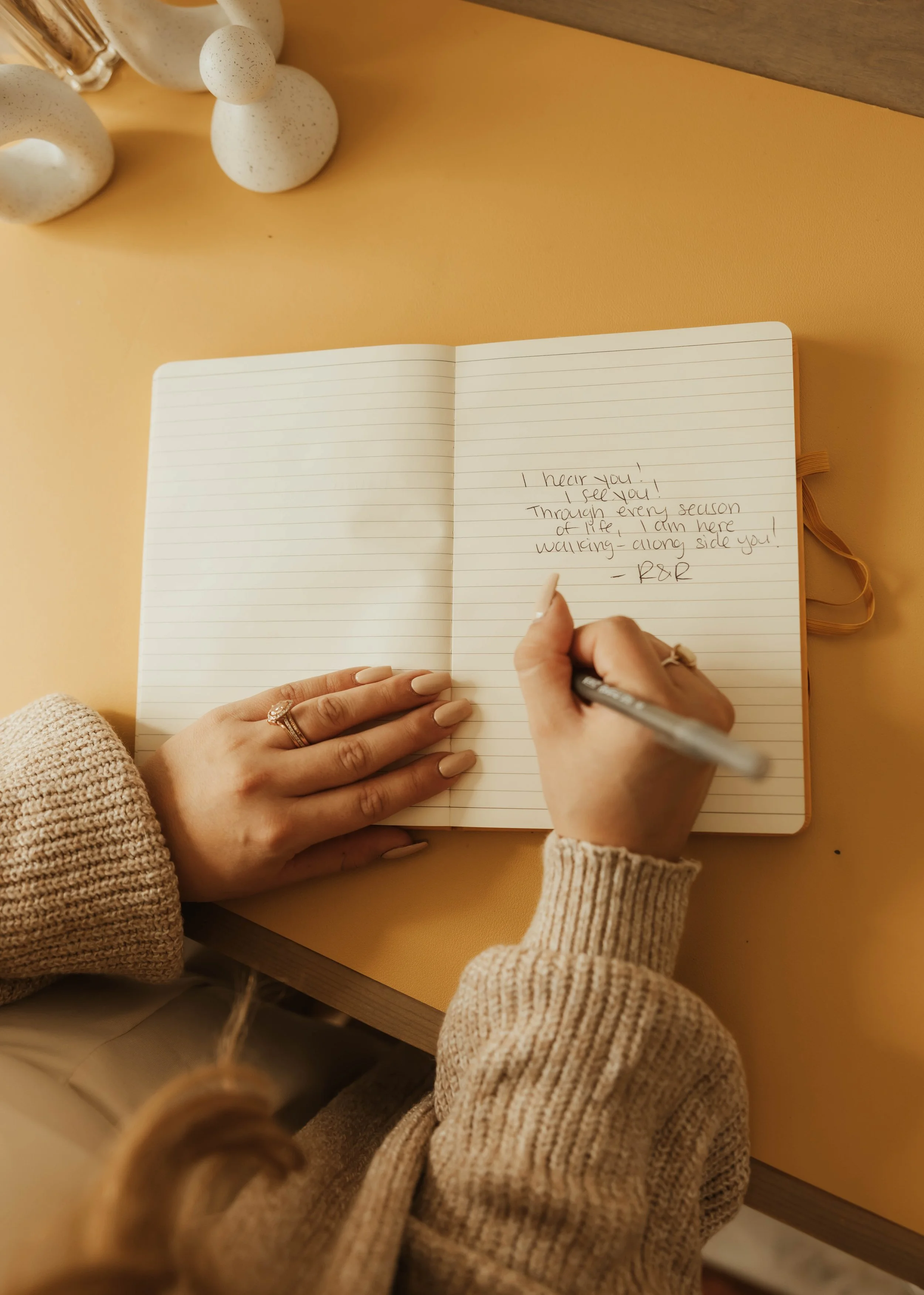 Person writing in a lined journal with a pen, on a yellow desk. Hands with rings touch the journal; the person wears a beige sweater. The journal has a handwritten message.