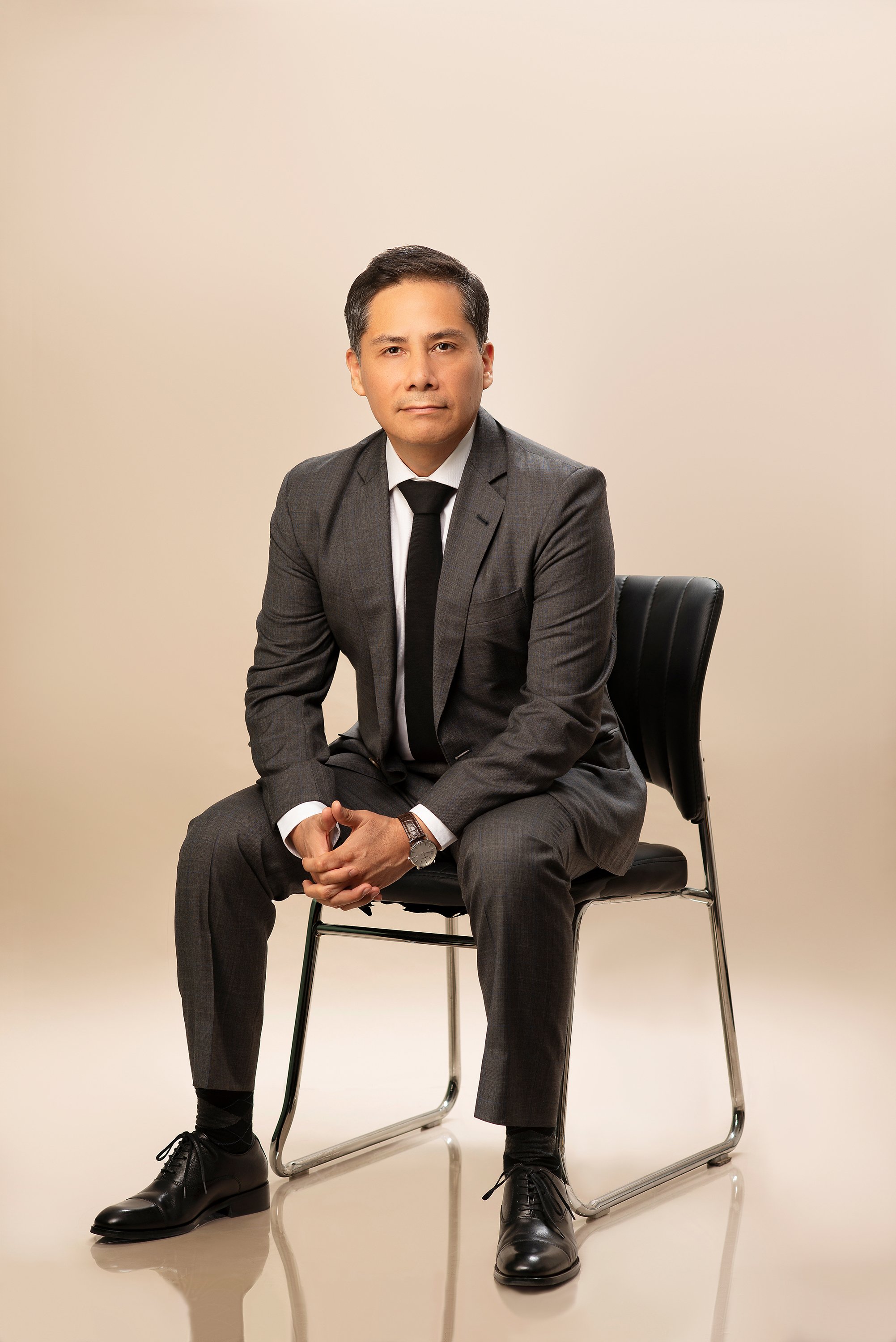 Retrato ejecutivo de cuerpo entero de hombre sentado con traje gris y corbata negra en estudio fotográfico.