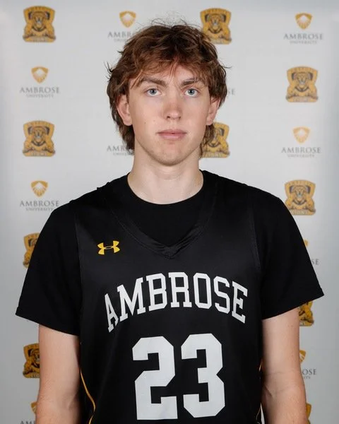 Young male athlete with curly red hair wearing a black basketball jersey with the number 23 and 'AMBROSE' written on it, standing in front of a backdrop with the Ambrose University logo.