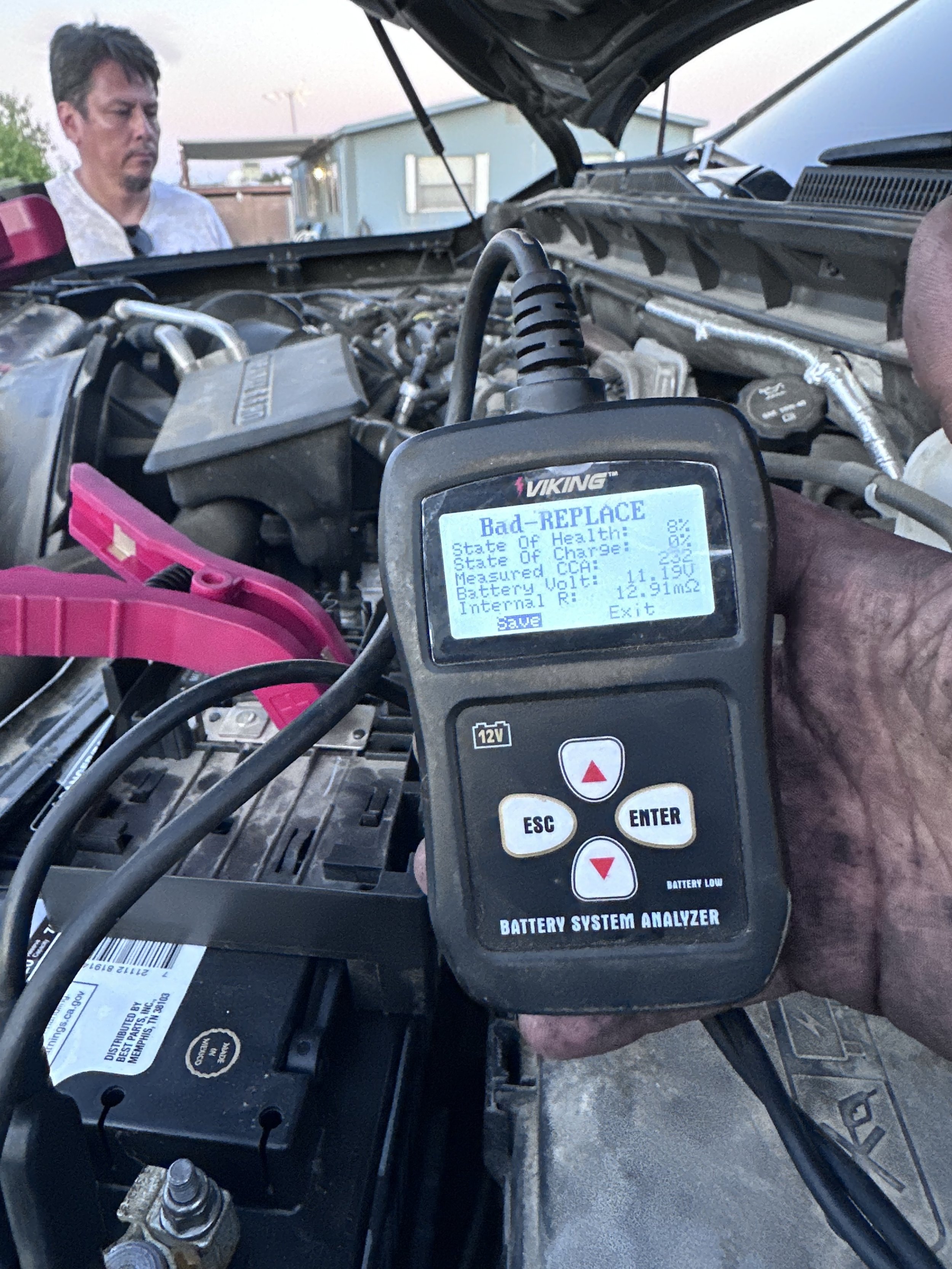 Close-up of a digital battery analyzer displaying a message about a bad battery, with a person inspecting the engine in the background and a mechanic or technician's hand holding the device.