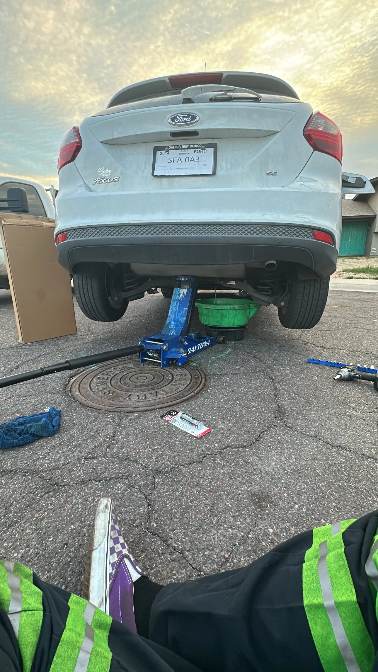 A person wearing a reflective safety vest and purple sneakers is sitting on the ground with legs stretched out, taking a photo of a white Ford Focus car that is raised on a blue hydraulic lift. The car's rear wheels are off the ground, and there are 
