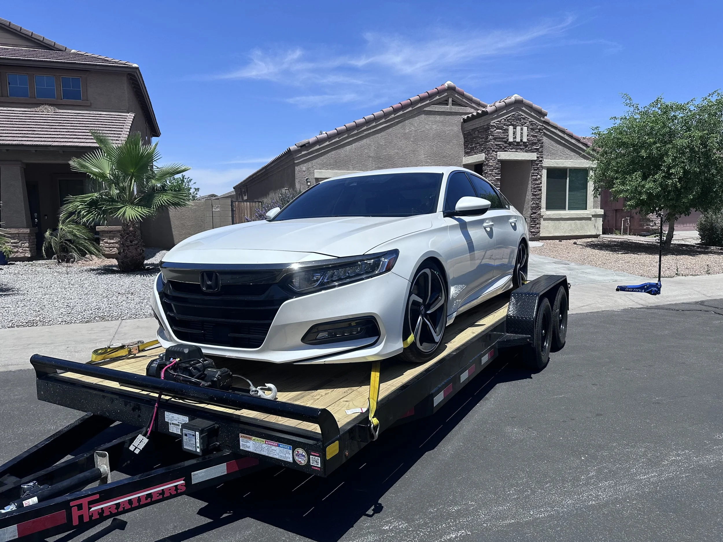White Honda sedan on a black flatbed trailer in front of a residential house with a stucco exterior and tiled roof, under a blue sky.