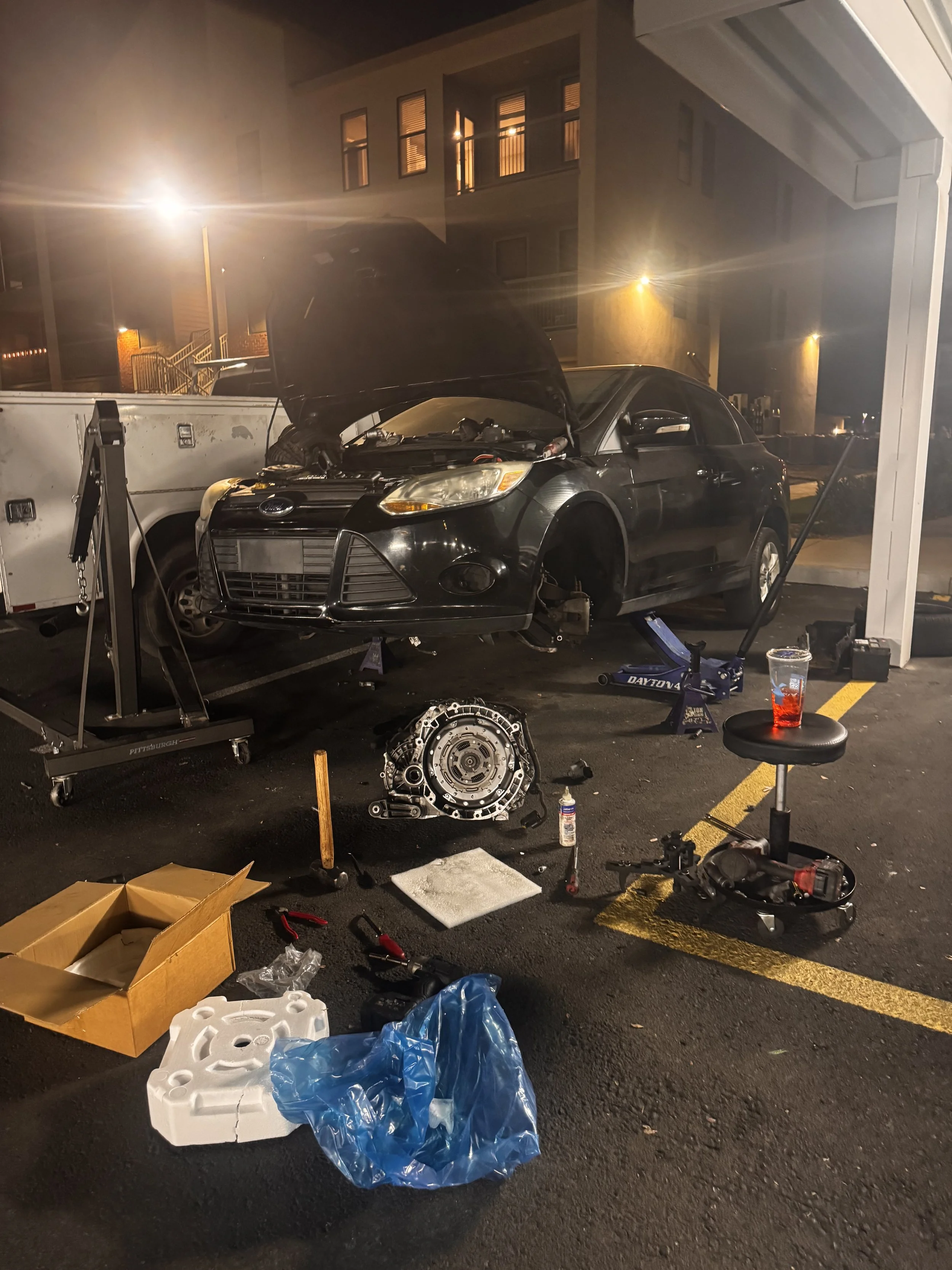 A black car with its hood open is parked in a parking lot at night. The front wheel is removed, and tools and car parts are scattered around, indicating car repair or maintenance.