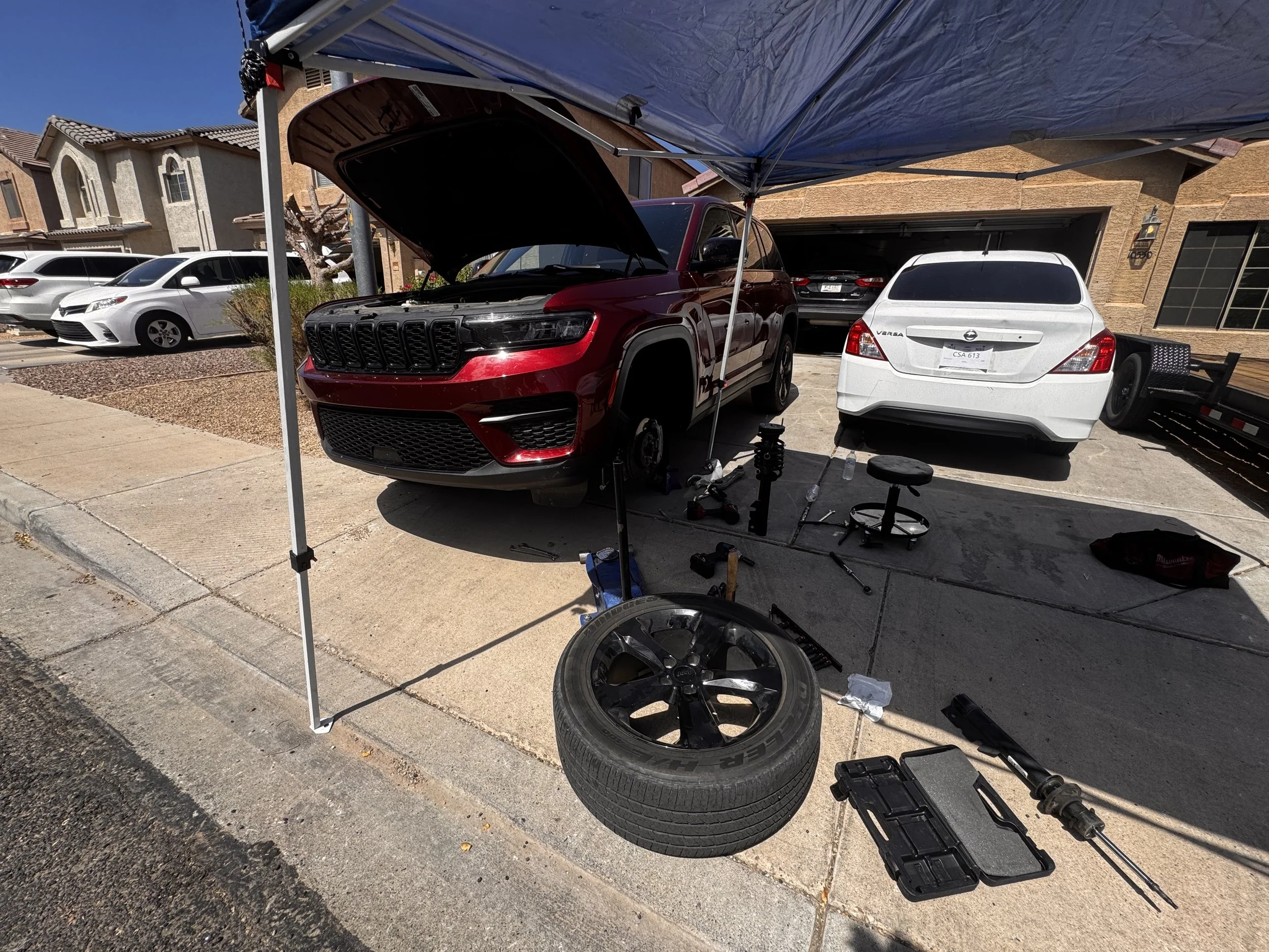 A red SUV with its hood open parked under a blue canopy in a residential driveway, with cars parked along the street and a house with yellow stucco exterior in the background.