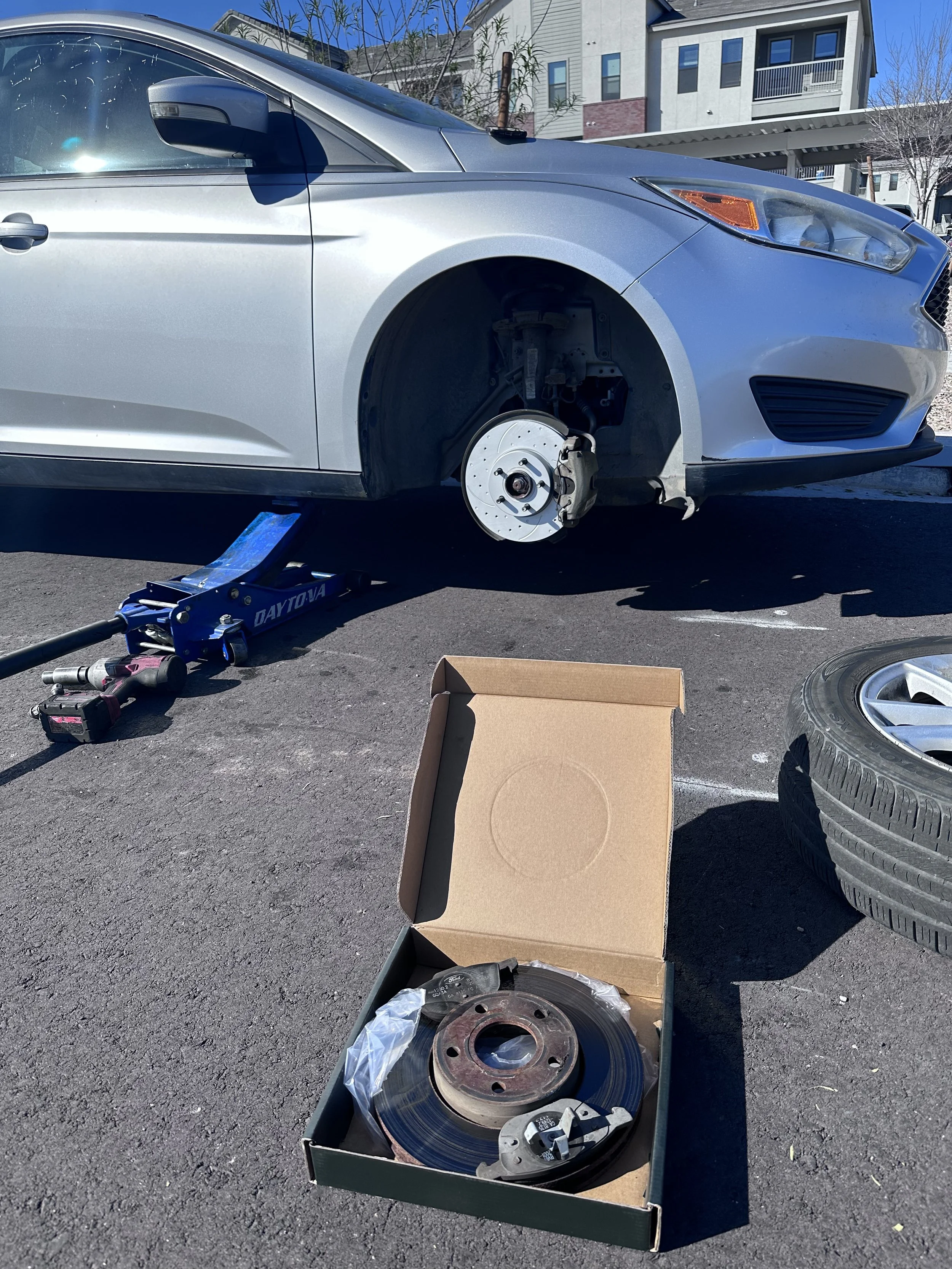 A silver car with the front right wheel removed, supported by a blue hydraulic jack, with an open cardboard box containing brake discs and other automotive parts nearby. The background shows a residential area with modern houses and a clear blue sky.
