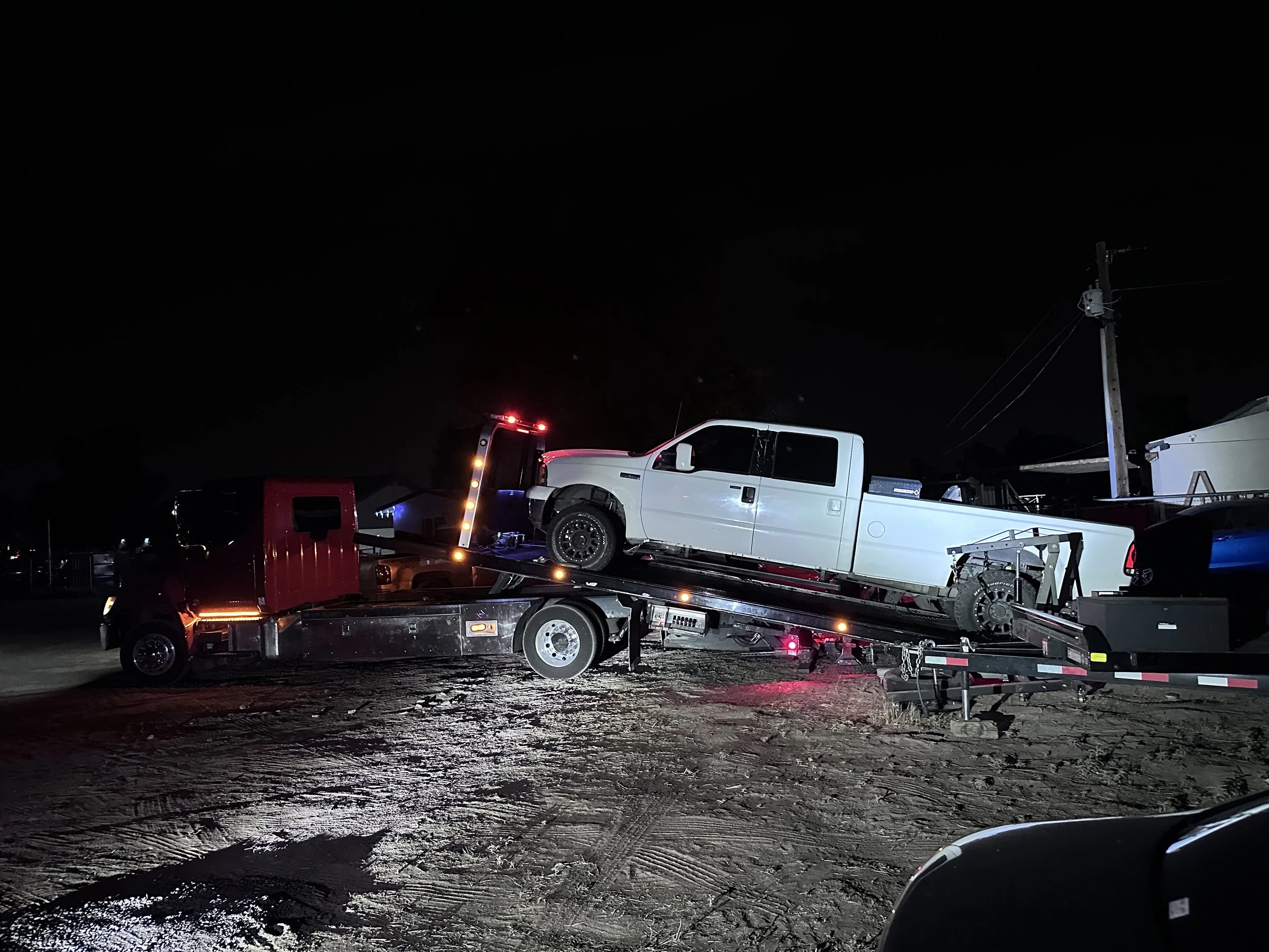 A white pickup truck loaded onto a flatbed tow truck at night, with a utility pole and power lines nearby, and a dark sky overhead.