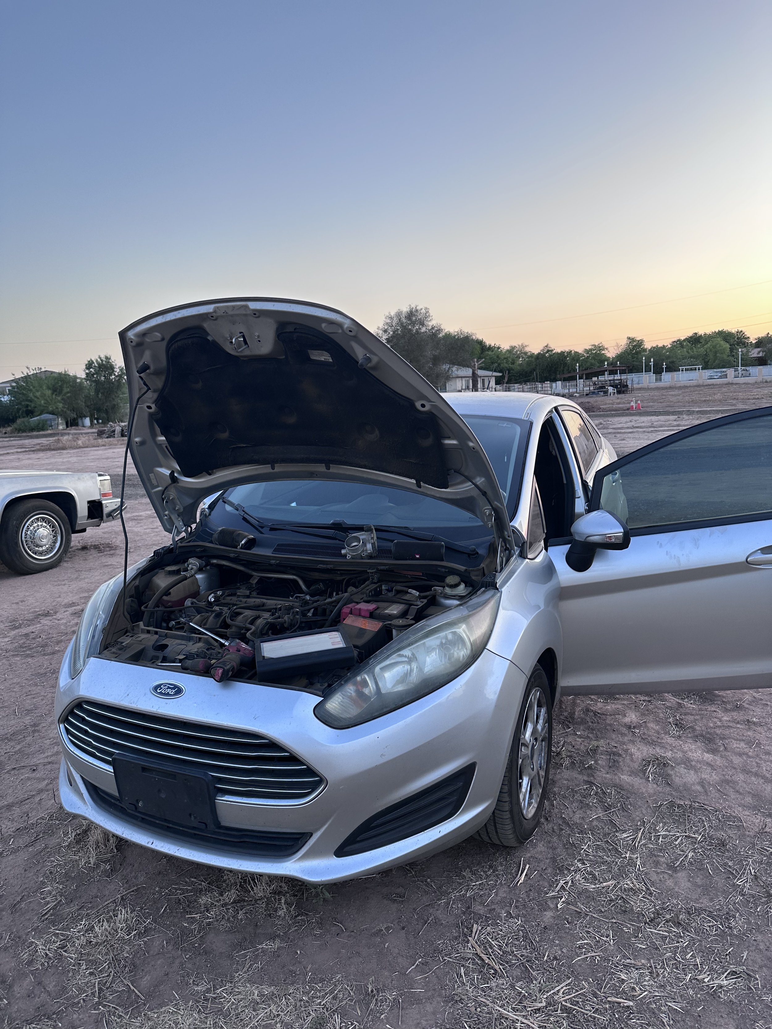 Silver Ford car with its hood open, parked on dirt ground, at sunset or sunrise with other vehicles and trees in the background.