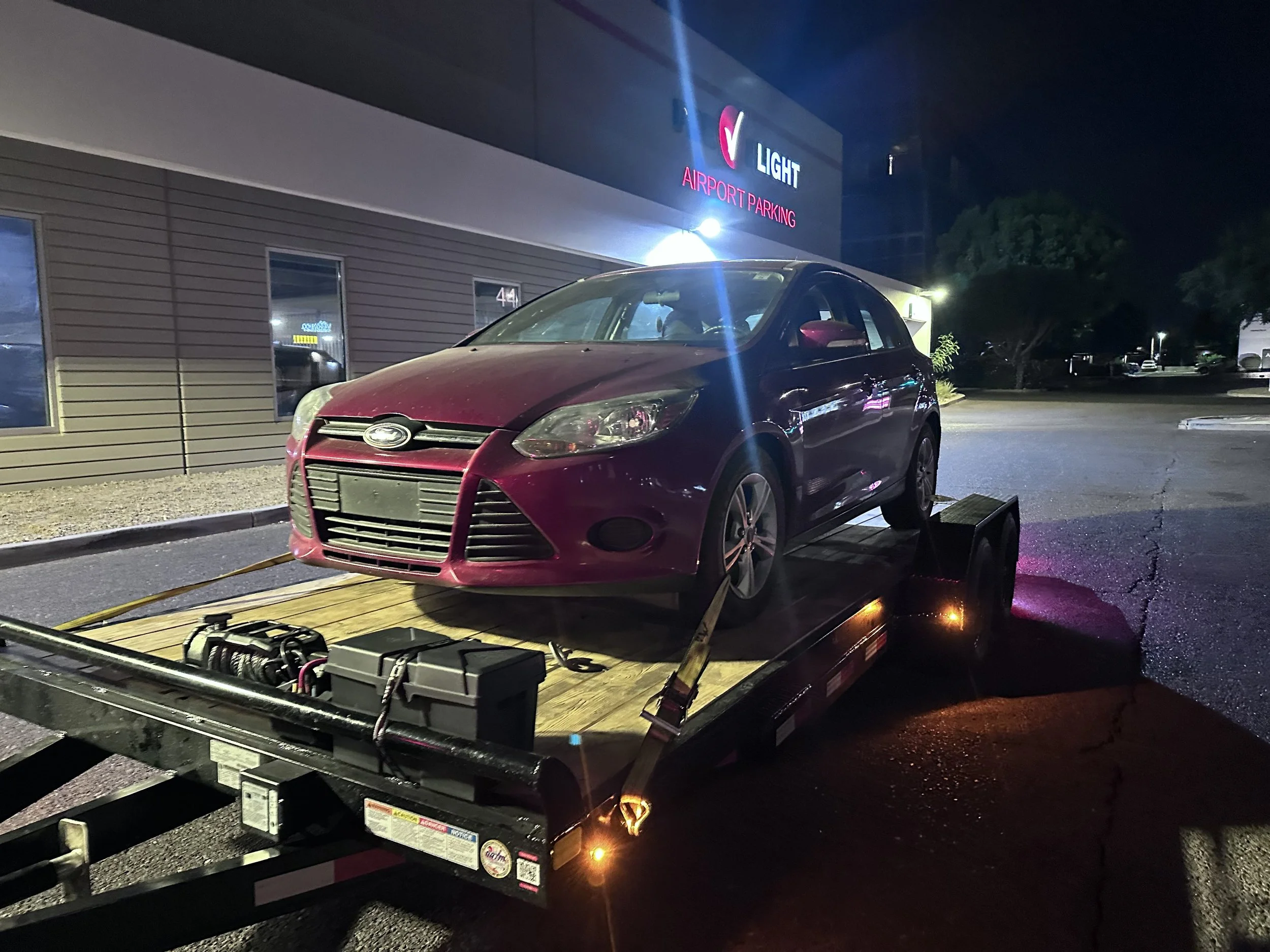 A red car on a flatbed trailer parked in front of a building with a sign that reads 'Light Airport Parking' at night.