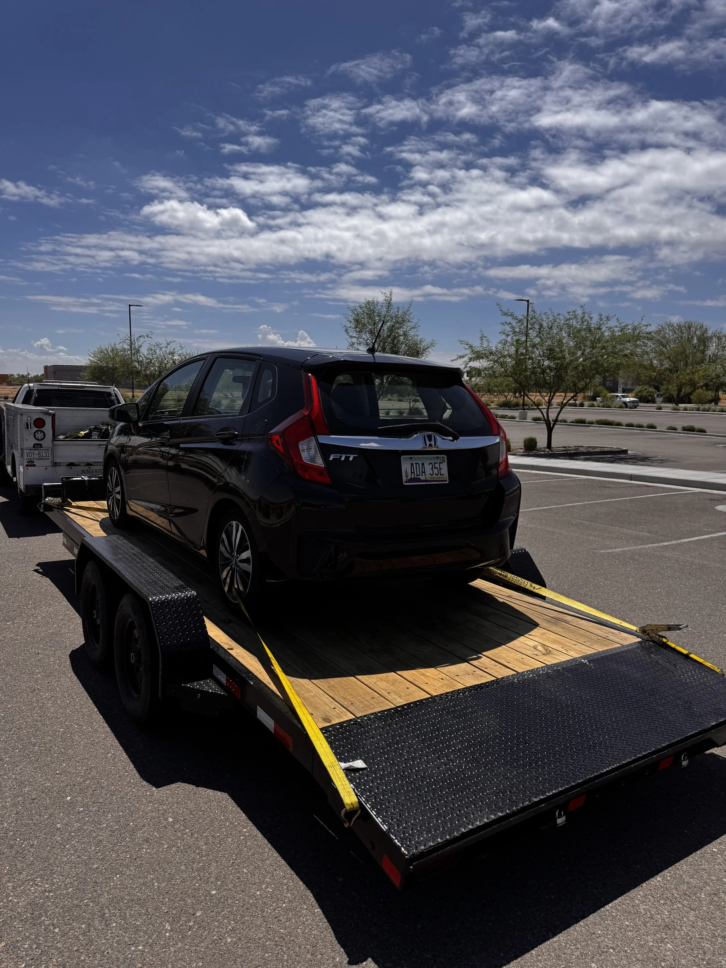 Black Honda Fit on a flatbed tow truck in a parking lot, with trees and streetlights under a partly cloudy sky.