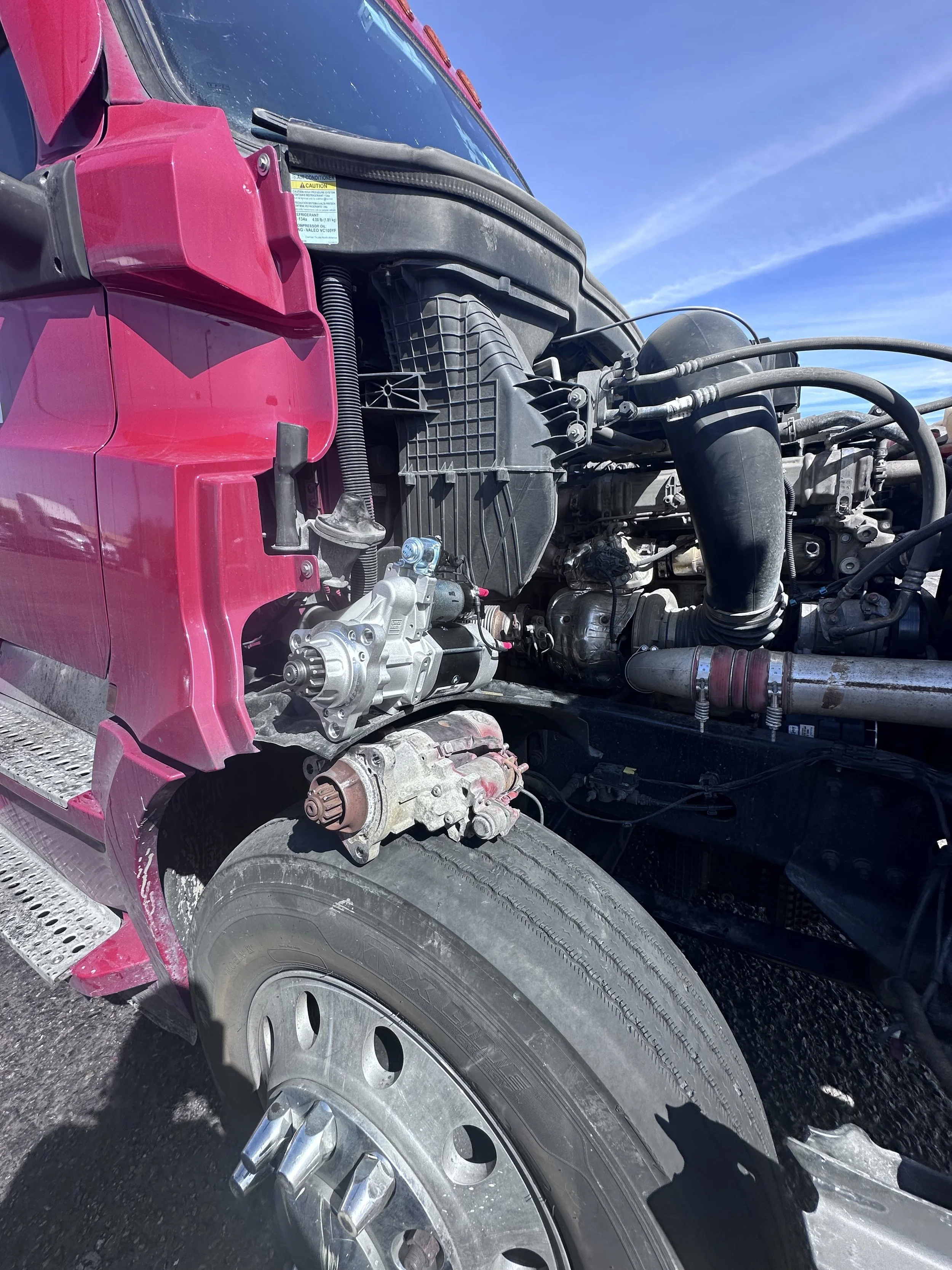 Close-up of a semi-truck with pink cab and exposed engine parts, including a large black air filter and assorted hoses, with a blue sky background.