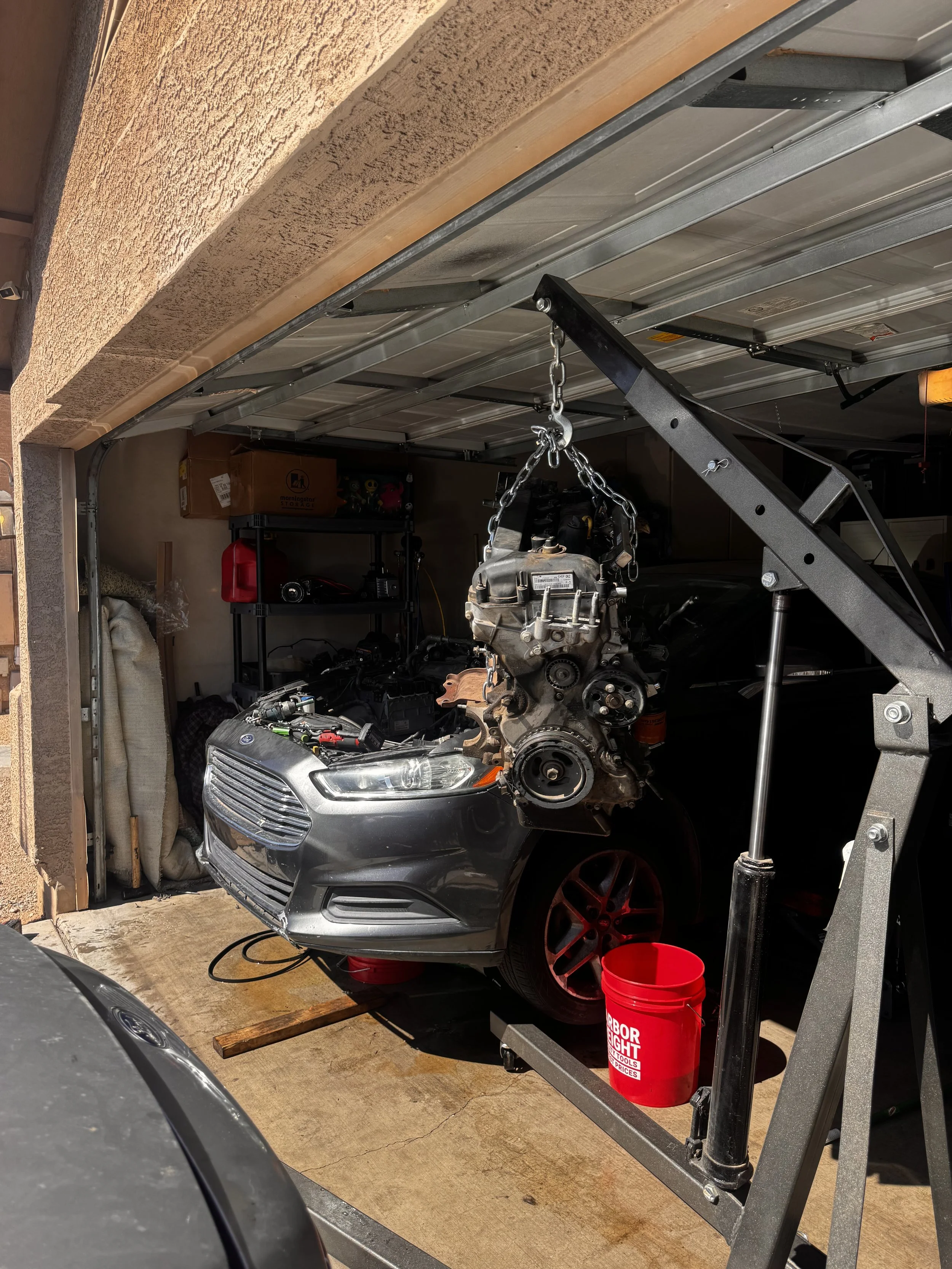 A gray car with a damaged front bumper on a concrete driveway. The car's hood is open, showing an engine hanging from a hoist next to it. There are tools and a red bucket nearby, and a garage in the background with shelves, storage boxes, and various