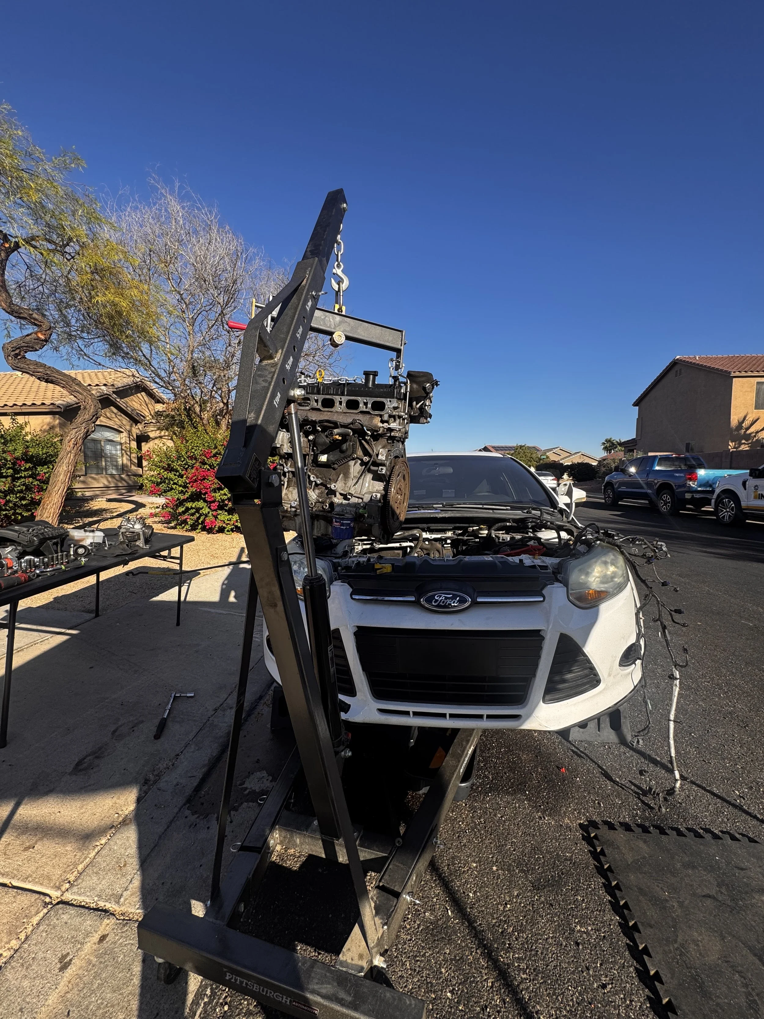 A white Ford car with its front bumper removed, placed on a black flatbed tow truck. The engine is being lifted from the car using a crane. Tools are scattered on a table to the left, and the scene is outdoors in a neighborhood with houses and trees 