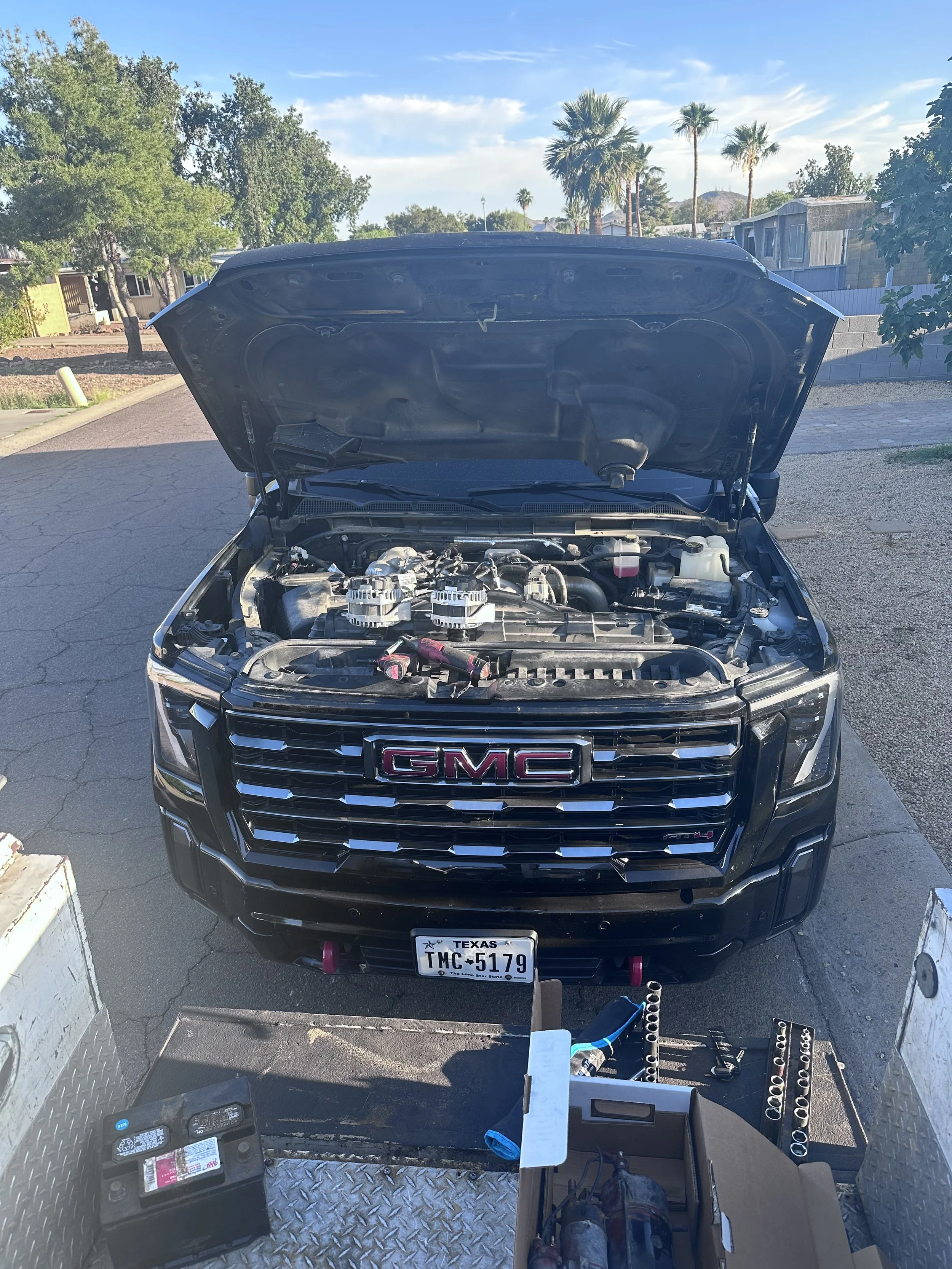 A black GMC vehicle with its hood open, positioned outdoors on a paved area, with tools and equipment in the foreground, including a cordless drill, socket wrench set, and a battery, indicating car repair or maintenance work.