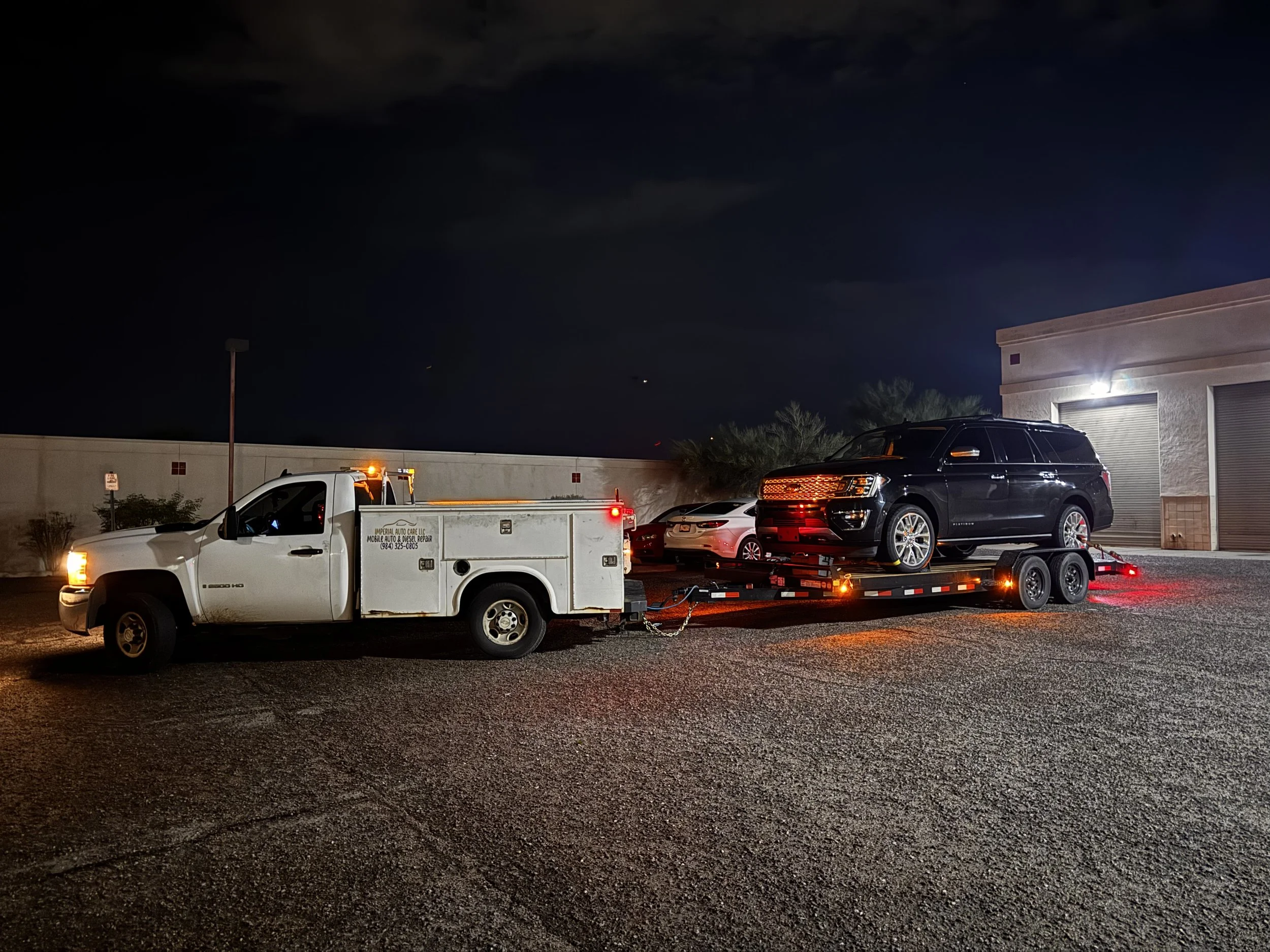 Nighttime scene in a parking lot with several parked vehicles, including a black SUV on a trailer, a white truck, and a white vehicle, with a building on the right side and a cloudy dark sky overhead.