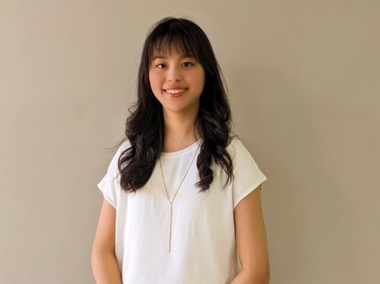 A young music teacher with long dark hair and a white shirt standing against a beige wall, smiling at the camera that can teach in Burnaby, Vancouver, New Westminster, and Richmond