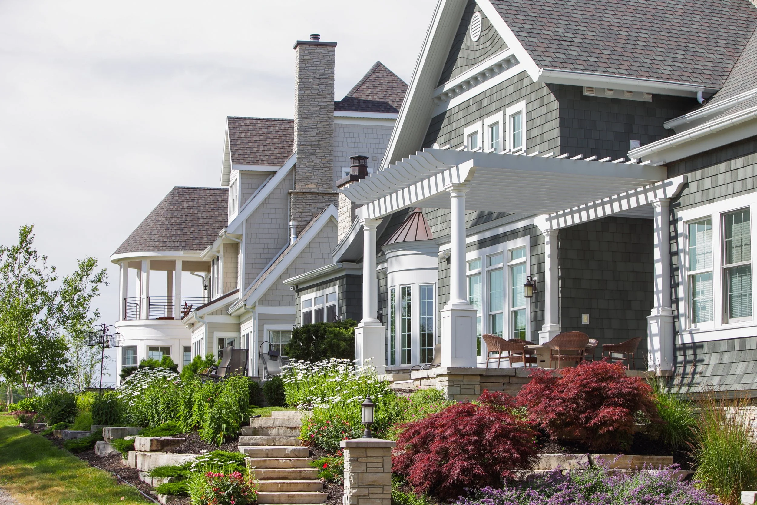 A row of large, multi-story houses with varied architectural styles and rooflines, featuring gray, beige, and dark green siding, with well-maintained gardens and a front porch with chairs and plants.