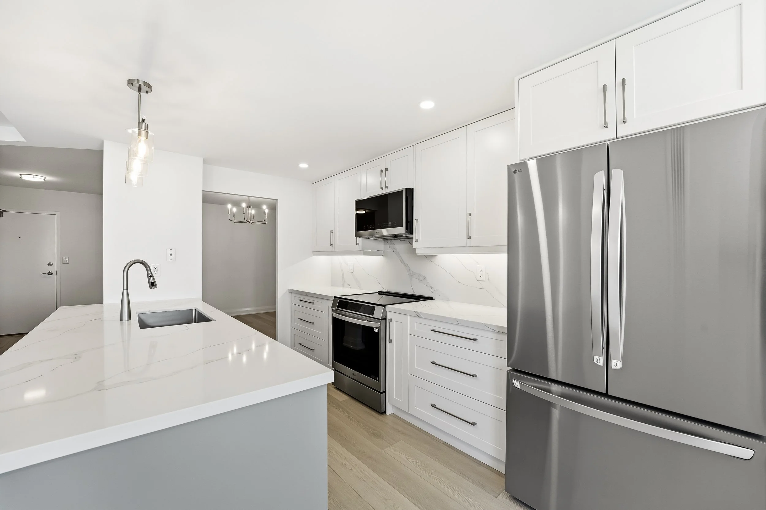 Modern white kitchen with stainless steel appliances, marble countertop island, pendant lighting, and hardwood floors.