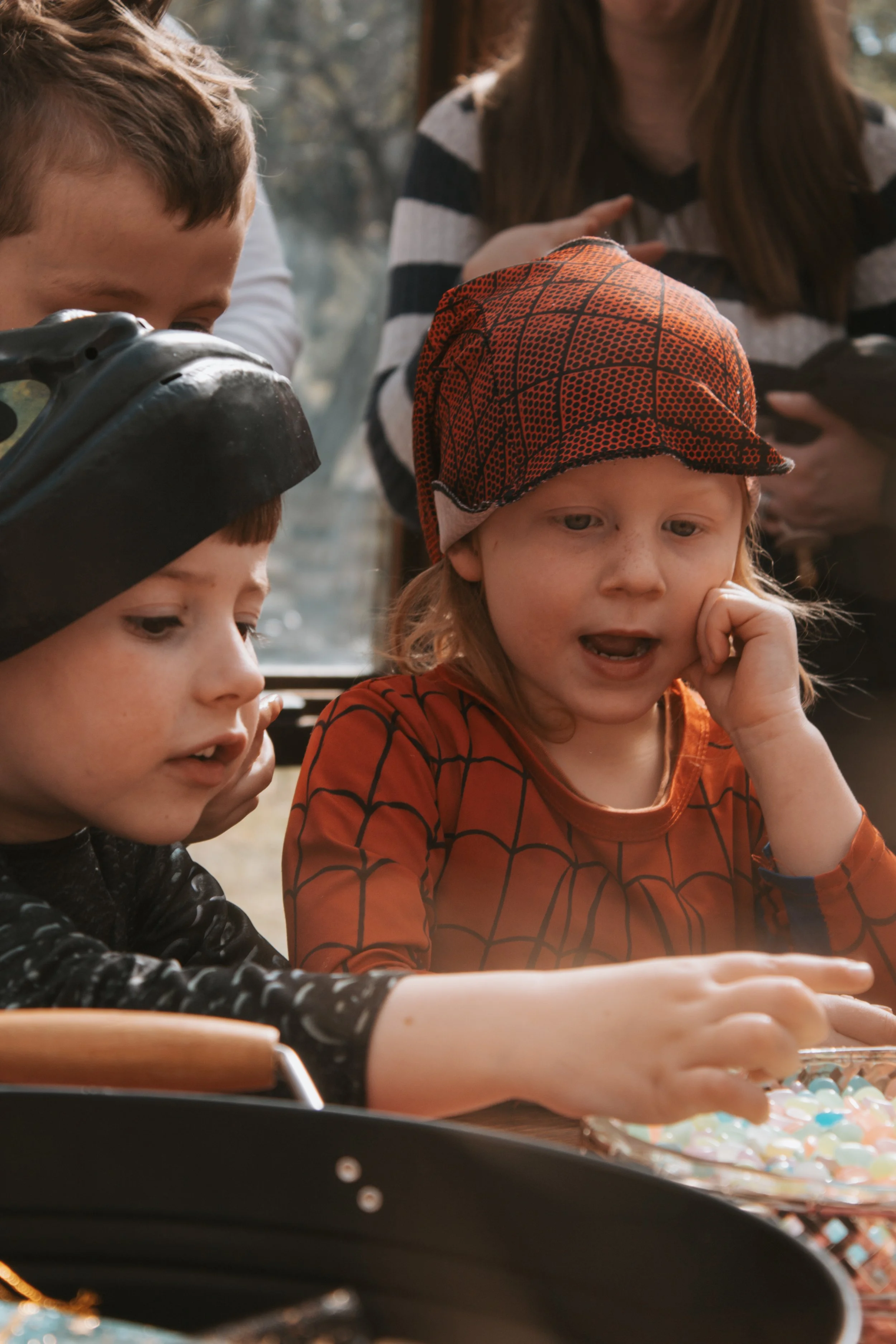 Children gathered around a table, looking at objects or candies, with an adult in the background.