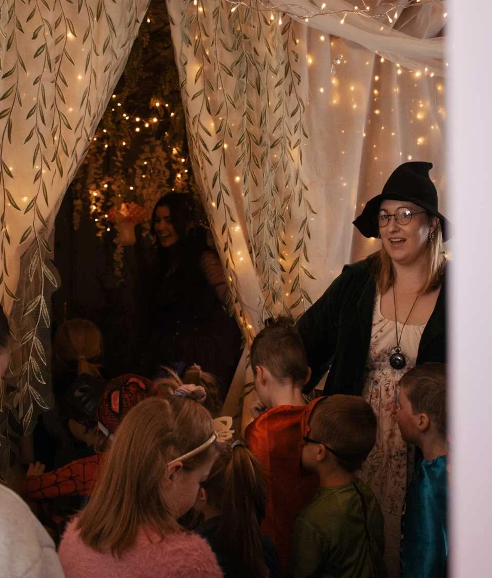 Children dressed in costumes gather around woman dressed as a witch, inside a decorated space with string lights and curtains.
