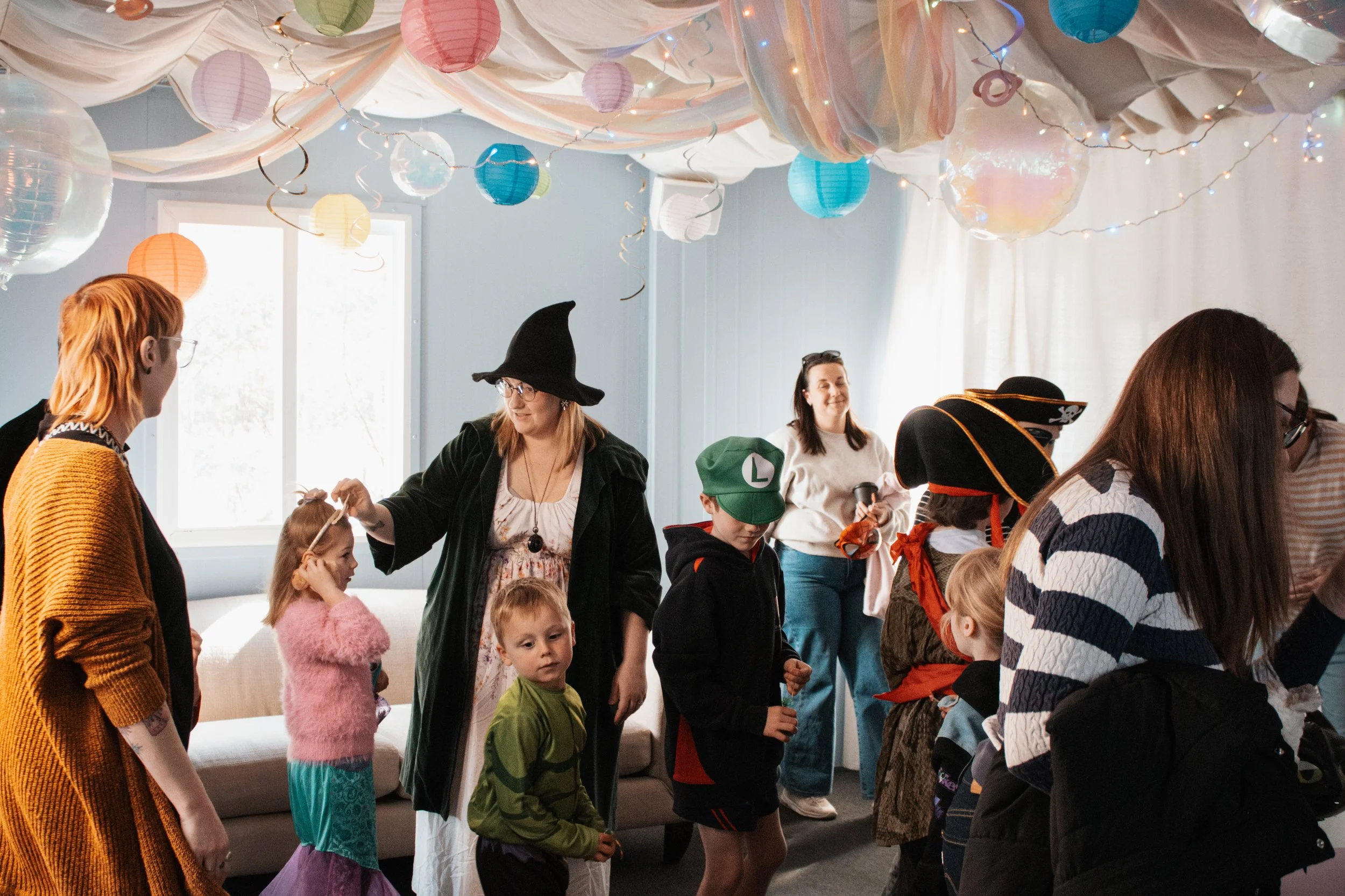 Children and adults in costume at a Halloween party, decorated with hanging paper lanterns and string lights.