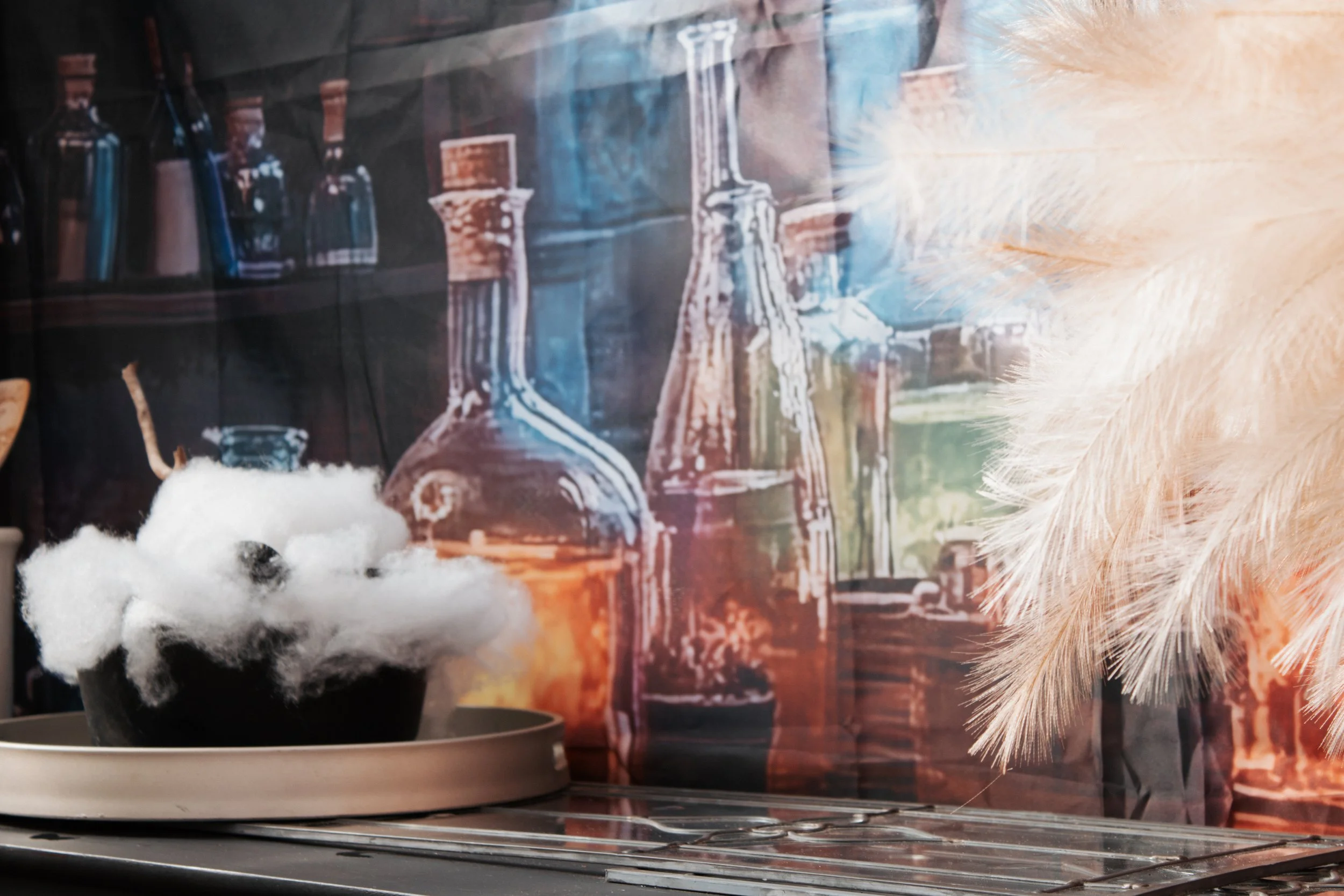 A small black bowl filled with white cotton balls, placed on a metal tray with a backdrop of a colorful, artistic wall mural featuring test tubes and bottles, and a branch of a light-colored artificial Christmas tree to the right.