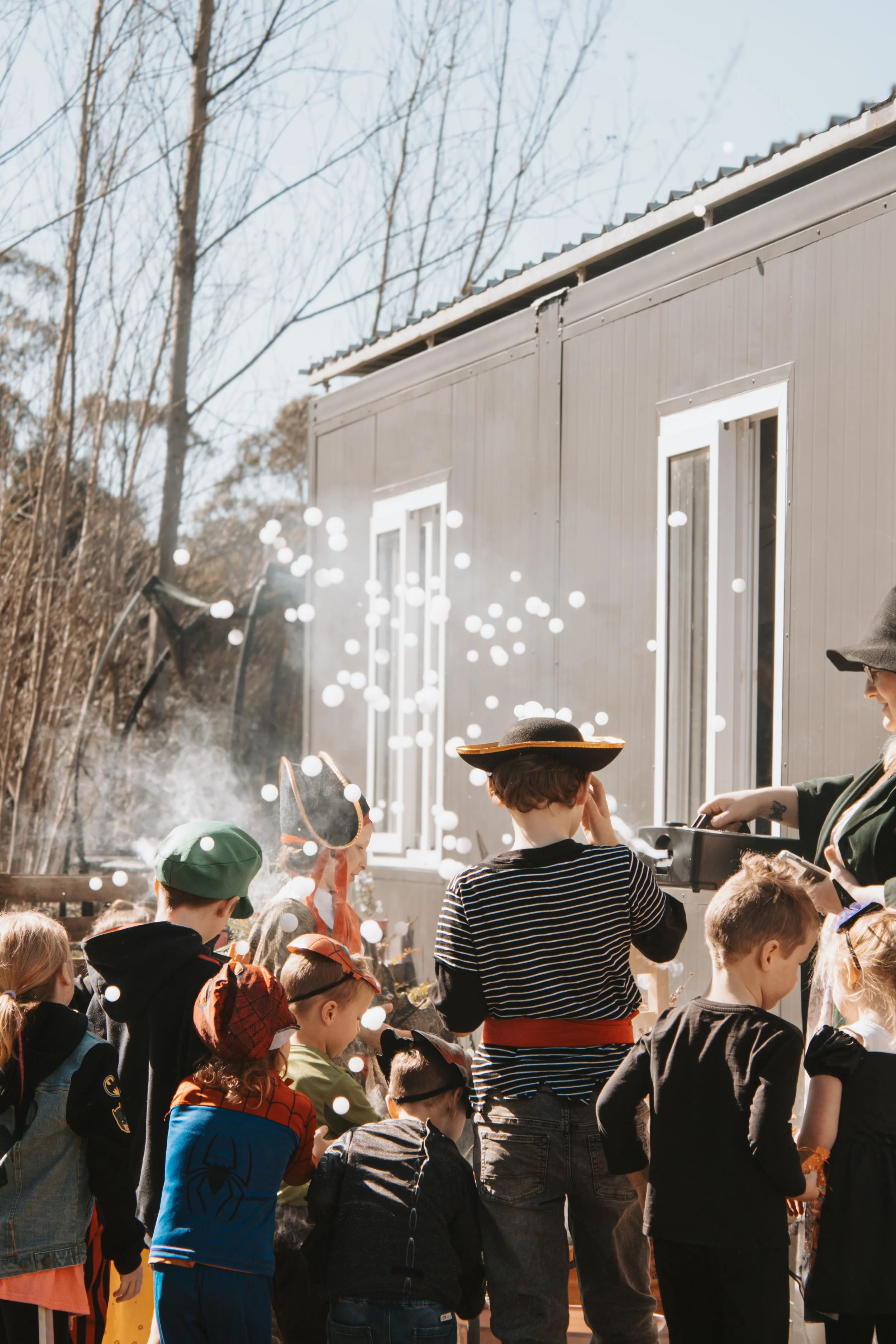 Children dressed in costumes gather outside near a gray building, as bubbles and steam fill the air during daytime.