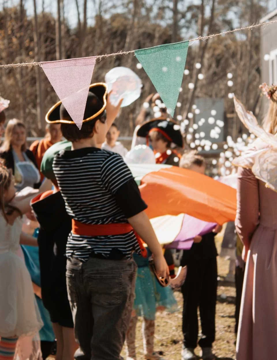 Children celebrating at a party outdoors, some wearing costumes and hats, with colorful bunting and falling confetti.