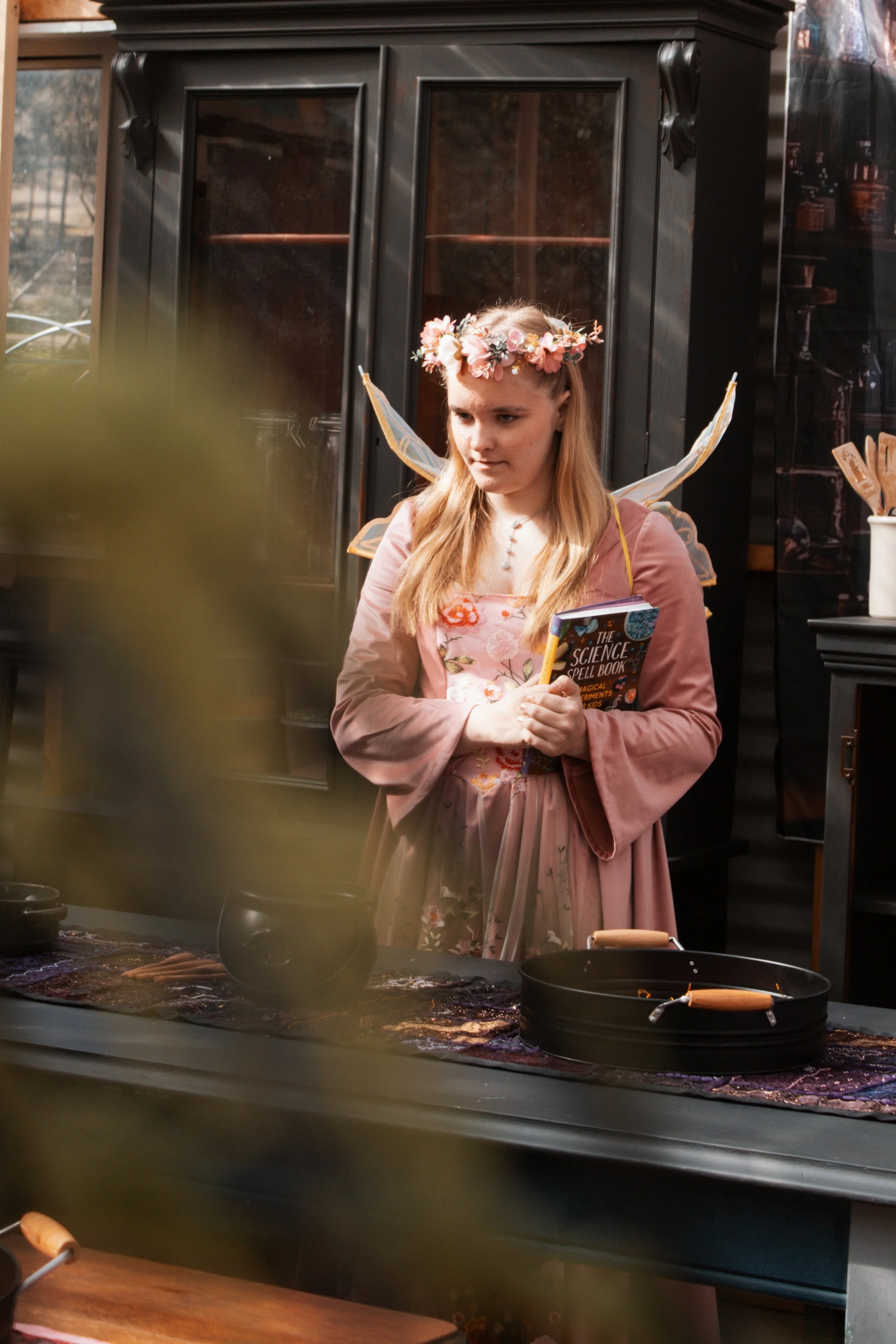 A young girl dressed as a fairy with wings, a floral crown, and holding a book titled 'The Science Spell Book' stands in a room with black furniture and plants, as she looks at a table with various objects.