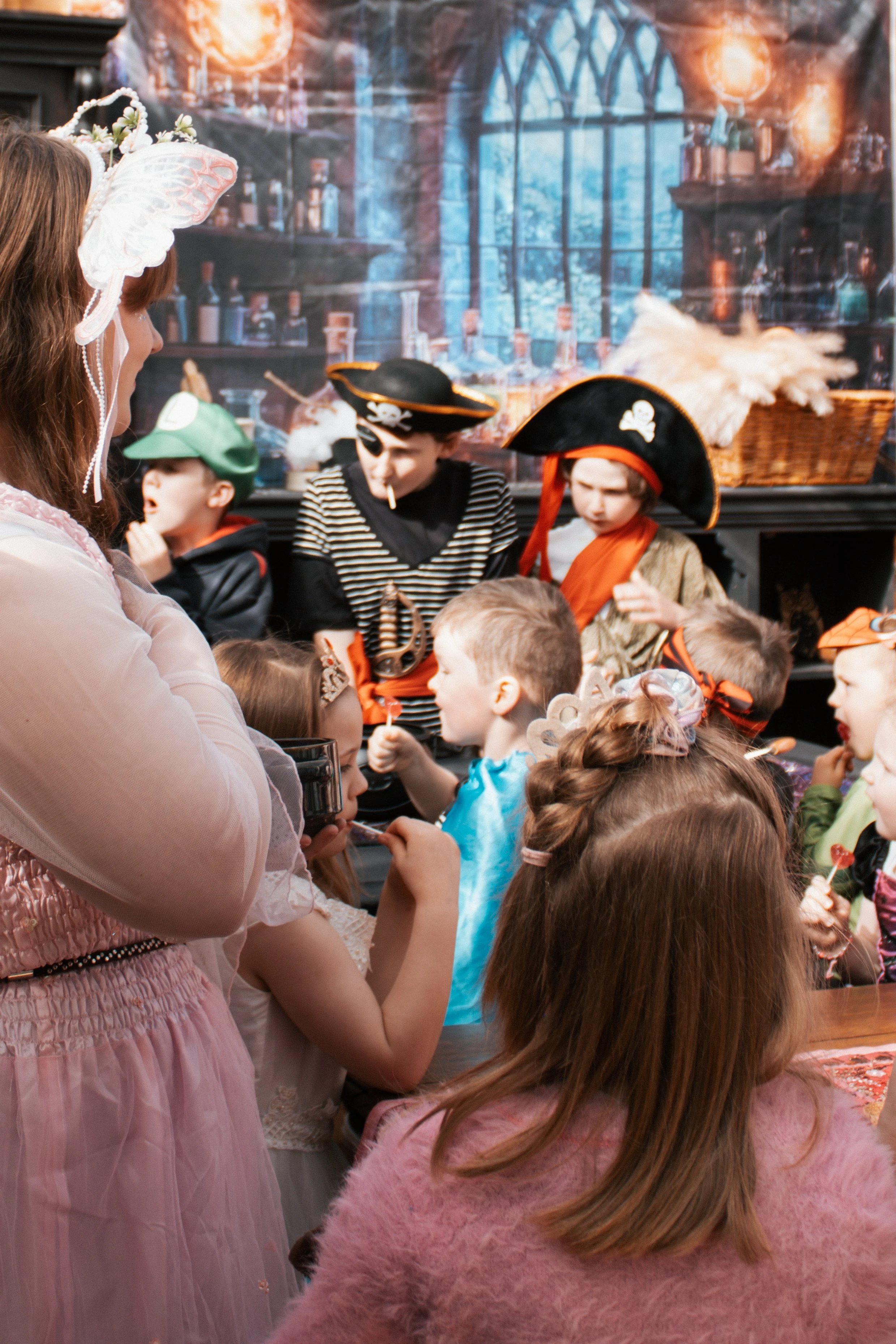 Children dressed in pirate costumes and fairy costumes gather around an adult at a birthday party, with pirate-themed decorations in the background.