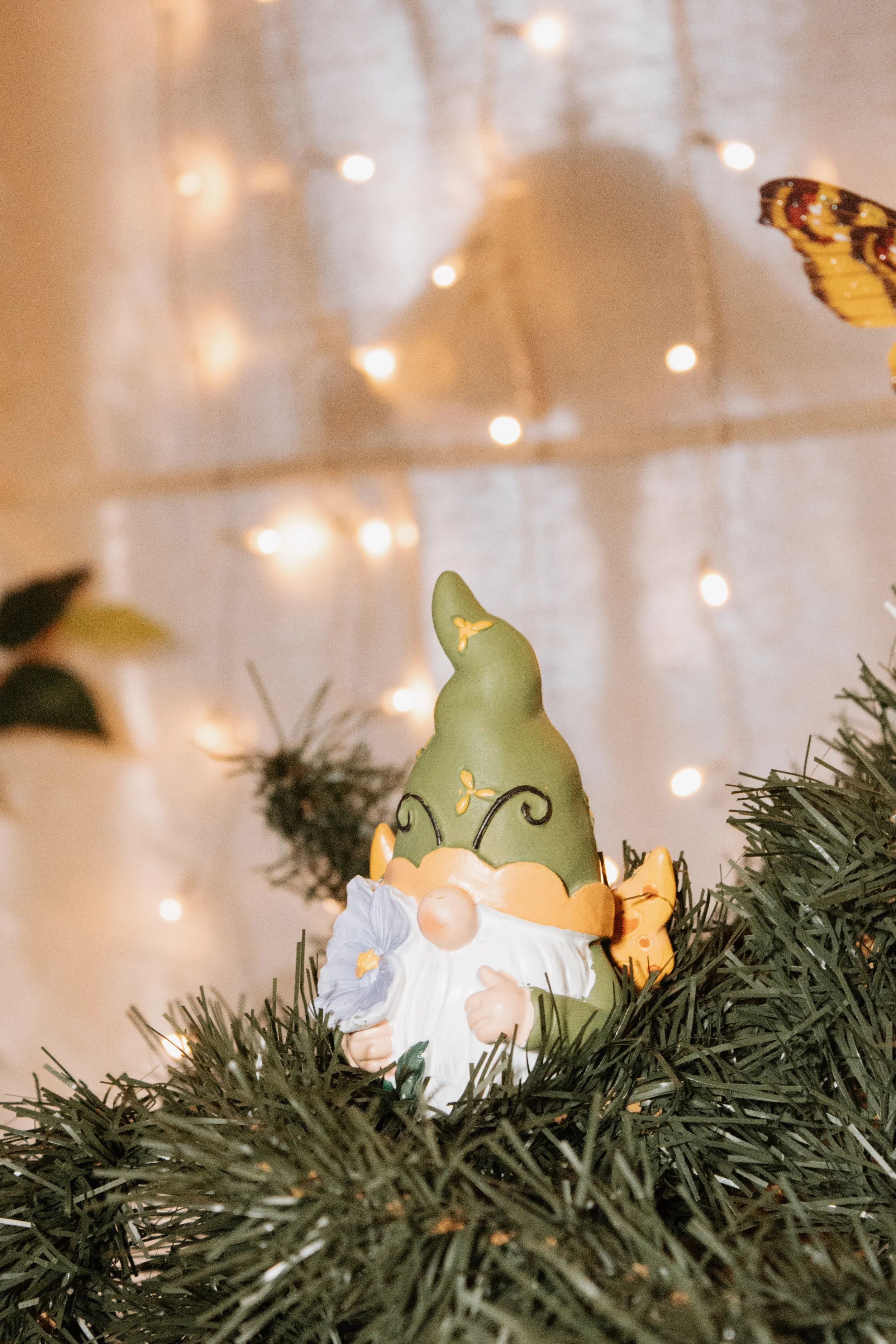 Decorative gnome ornament on Christmas tree surrounded by green pine branches, with a blurred background of warm fairy lights and a butterfly.