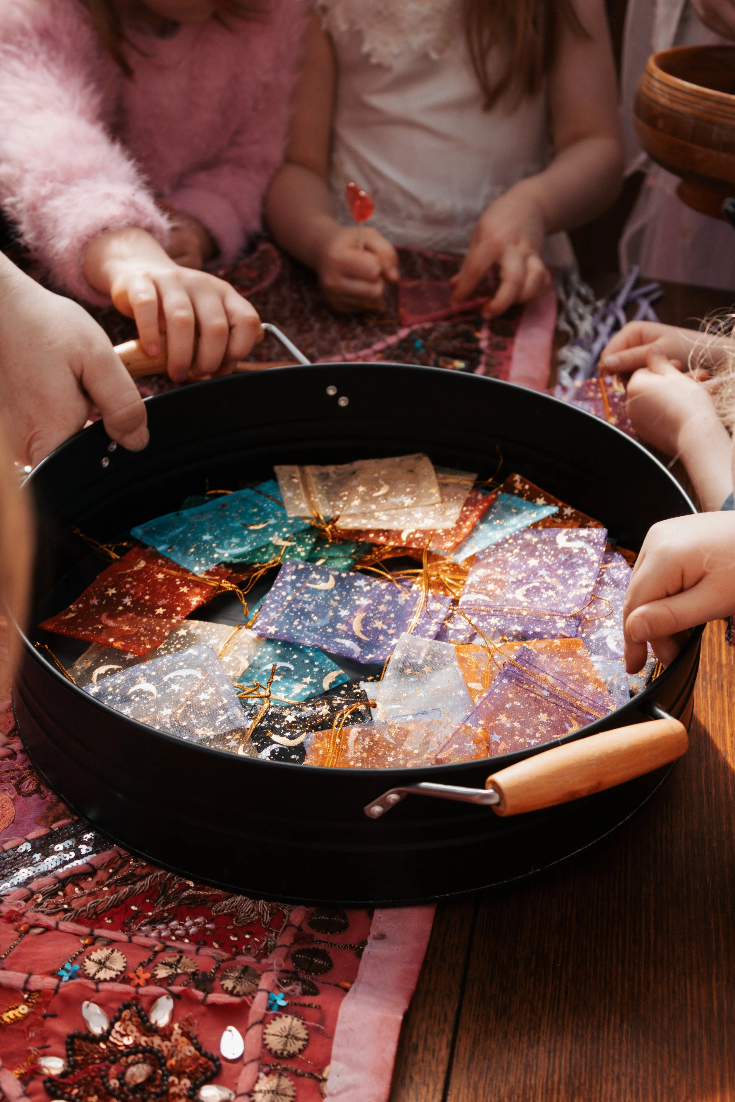 Children gathered around a black tray filled with colorful, glittery star and moon-themed fabric pieces, possibly for craft or decoration.